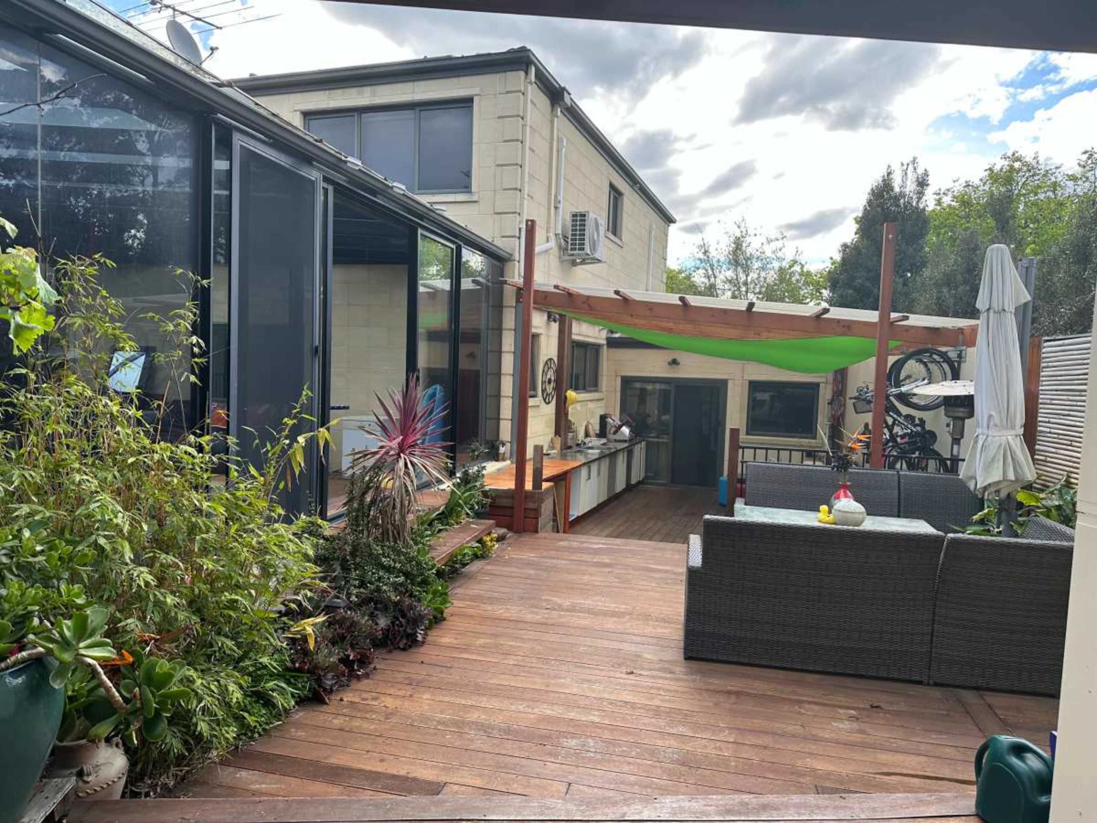 The image shows a wooden deck area with outdoor seating and a kitchen space in the background, surrounded by greenery and under an overcast sky.