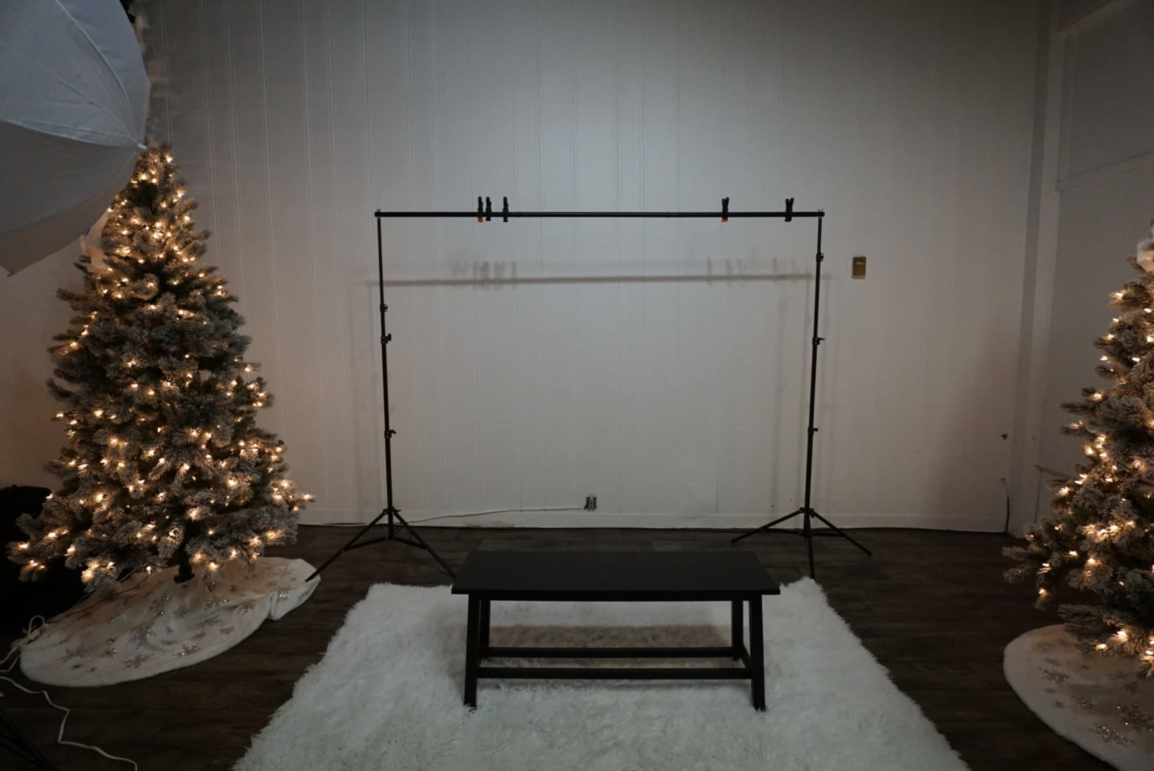 A minimalist studio setup with a black table in front of a backdrop frame, flanked by two lit Christmas trees on either side.