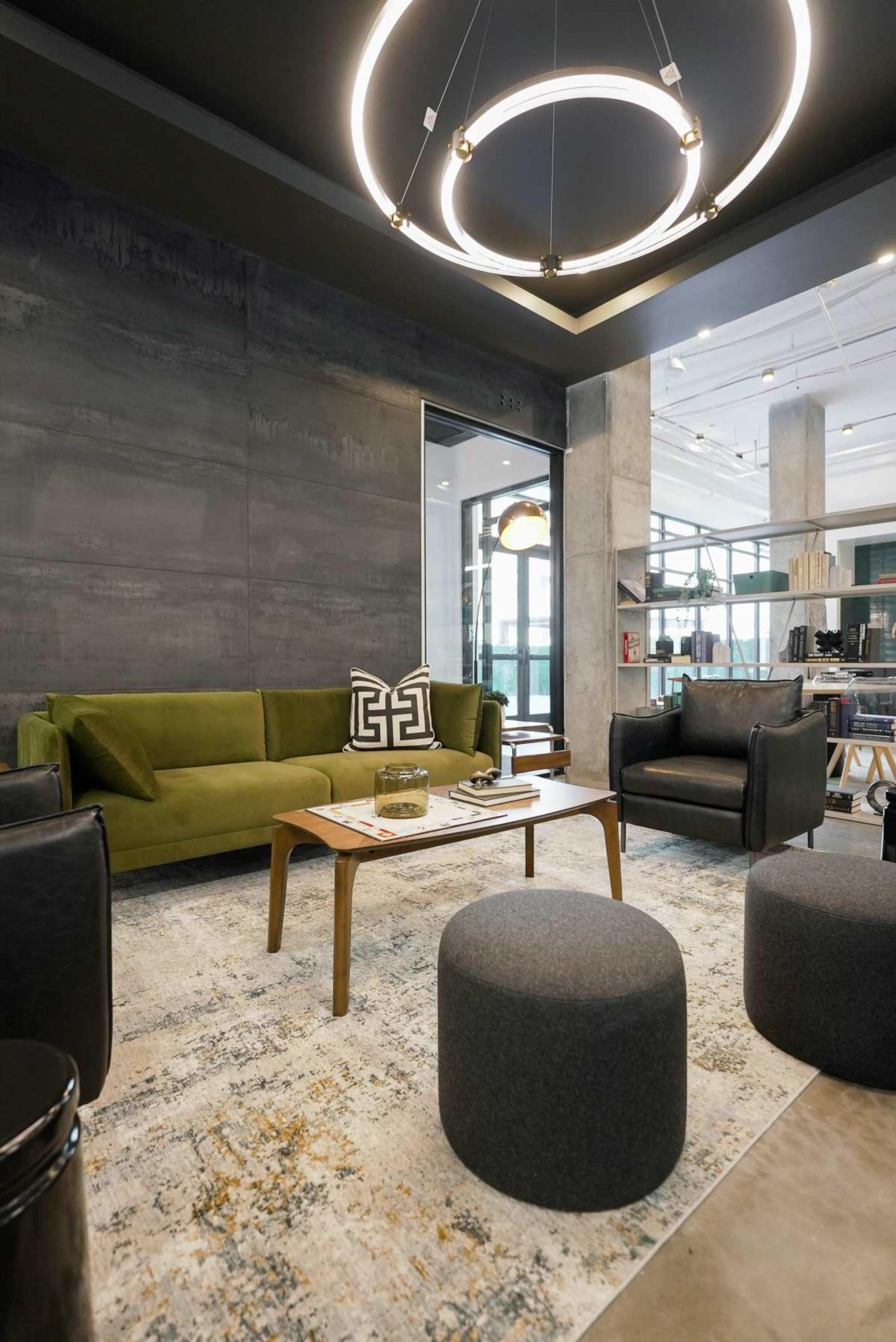 A modern lounge area featuring a green sofa, two black armchairs, a round wooden table, and several gray poufs on a patterned rug under a large circular chandelier.