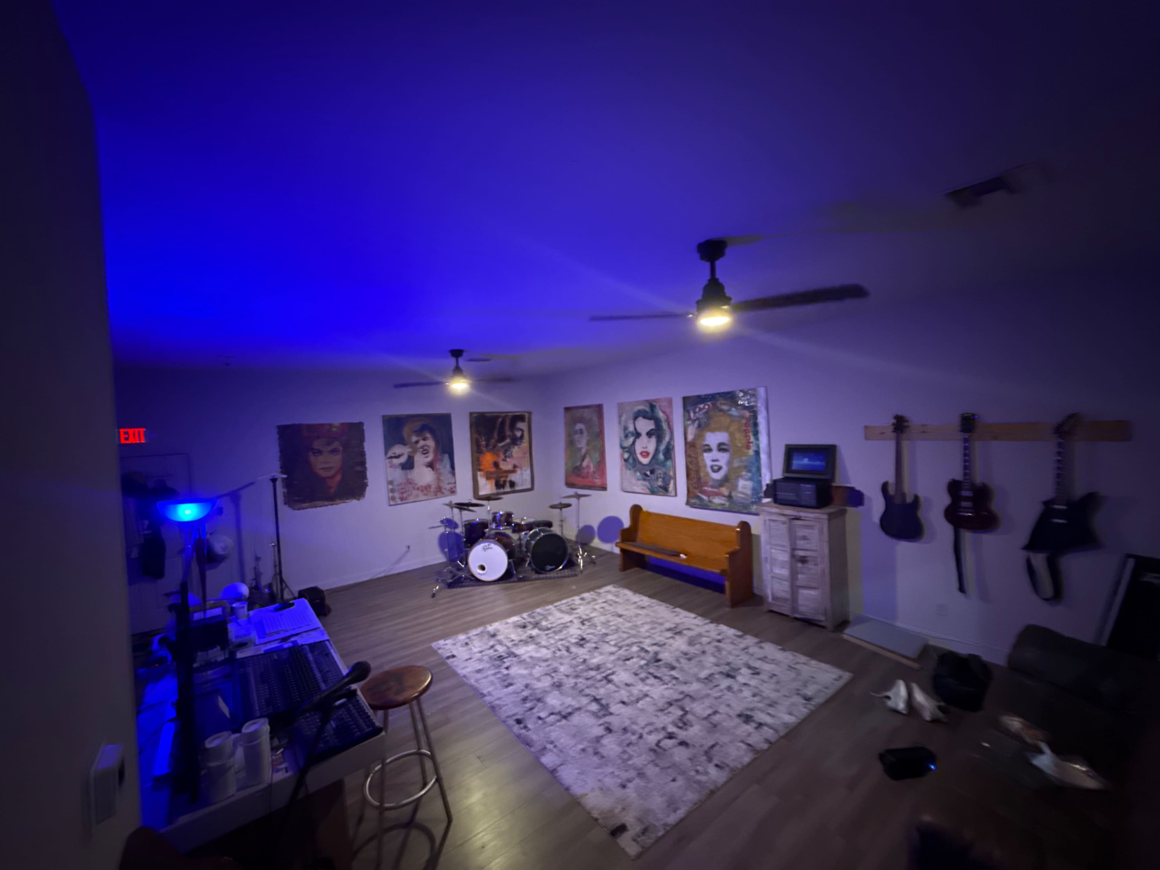 The image shows a dimly lit room featuring a drum set, several guitars hanging on the wall, and colorful artwork of faces.