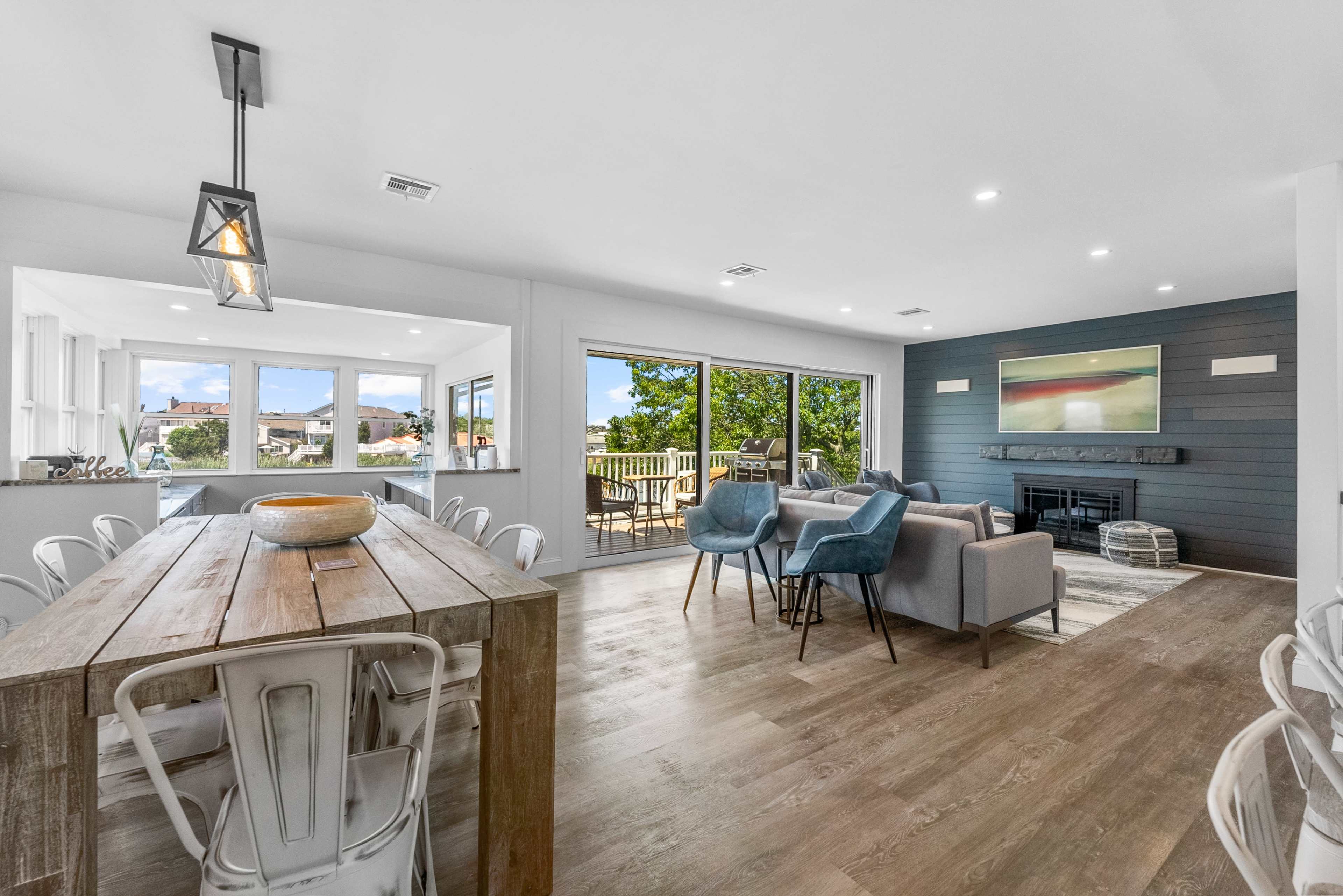 The image shows a modern open-concept living and dining area with large windows, a wooden dining table, and a cozy seating arrangement beside a fireplace.