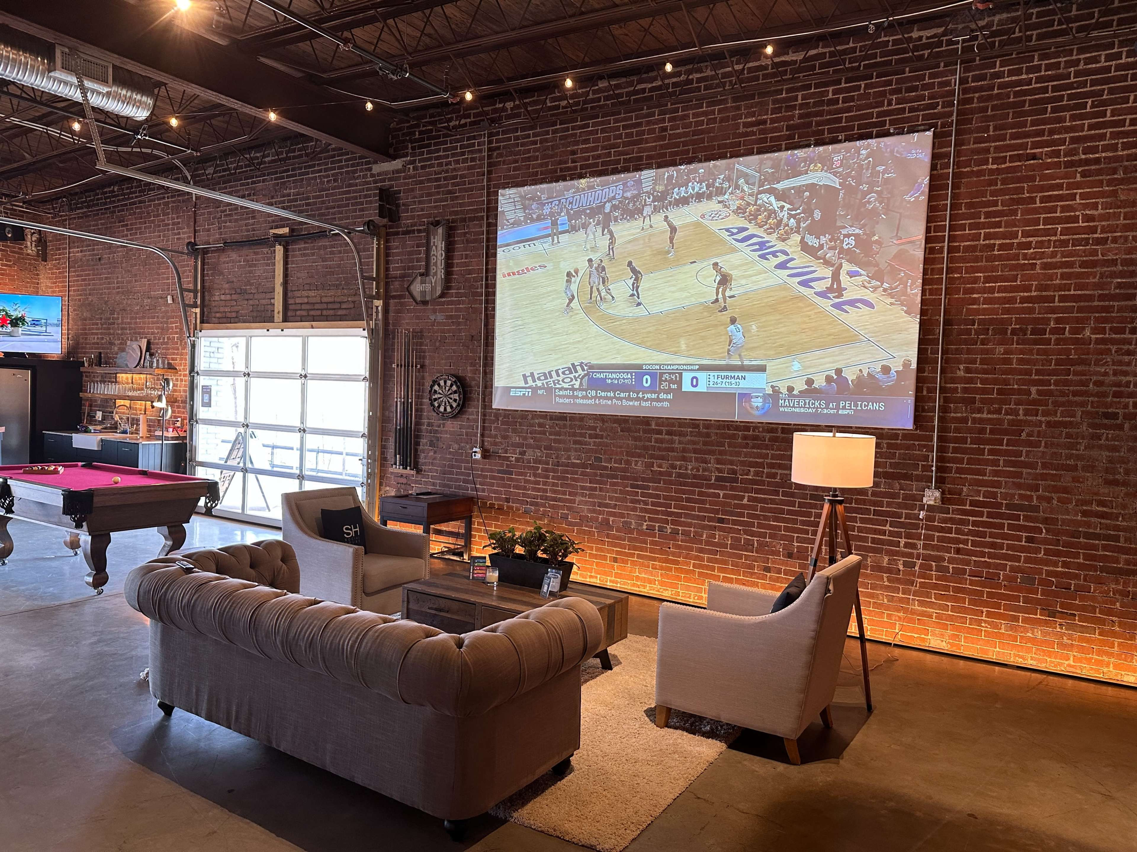 The scene features a stylish lounge area with a large screen displaying a basketball game, a pool table, and a brick wall backdrop.