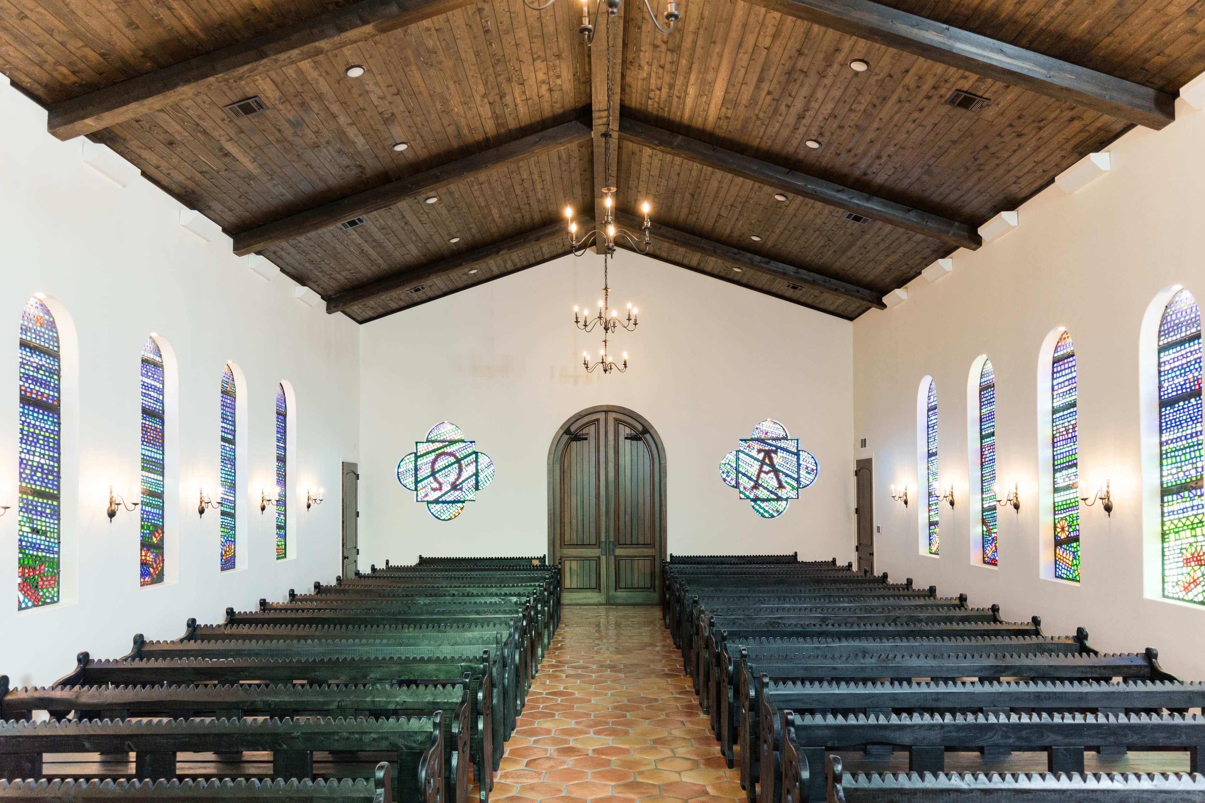 The interior of a church features rows of black wooden pews, colorful stained glass windows, and a wooden beam ceiling.