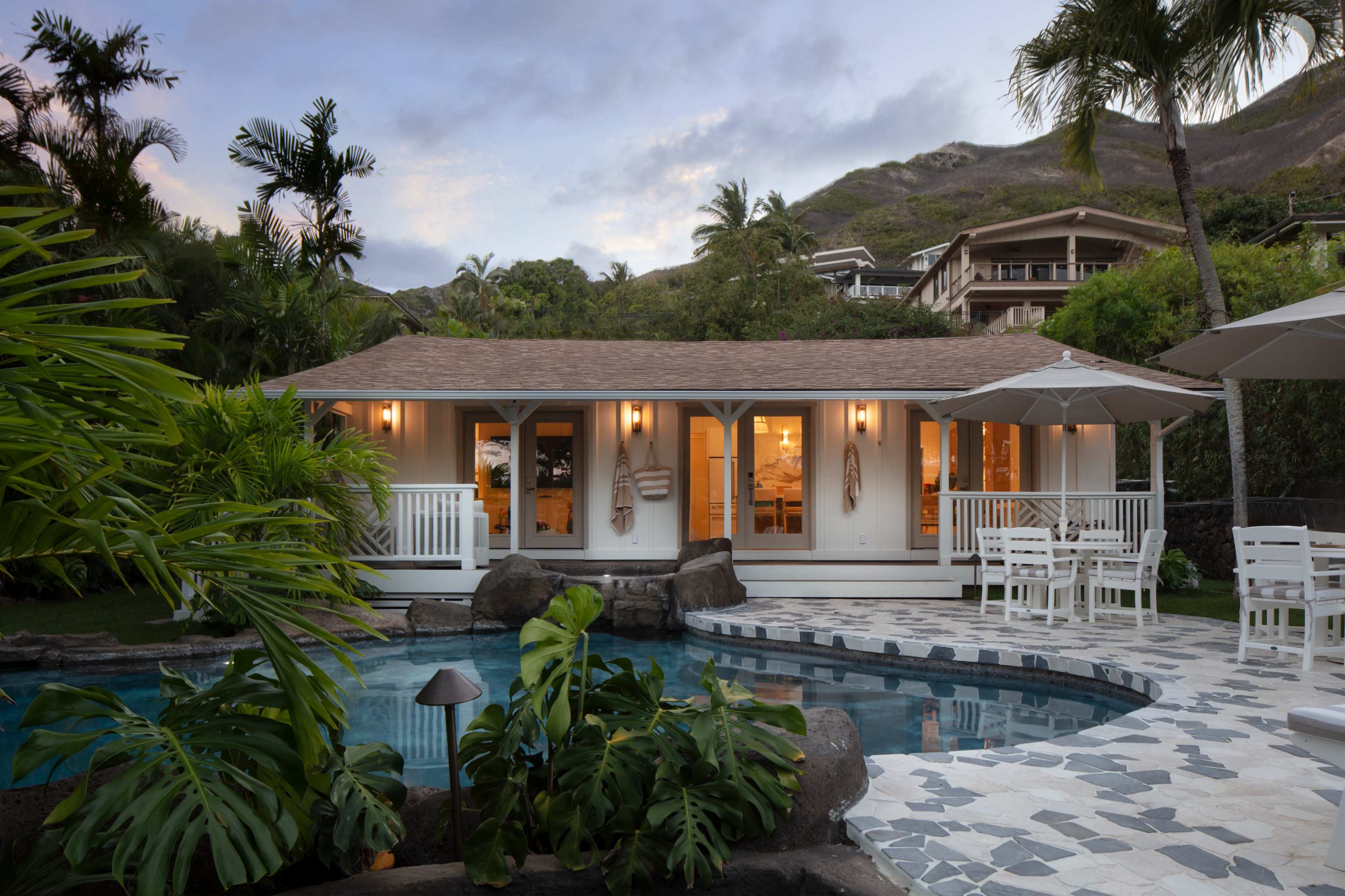 A modern cottage with large windows sits beside a pool surrounded by stone and tropical landscaping, with mountains in the background.