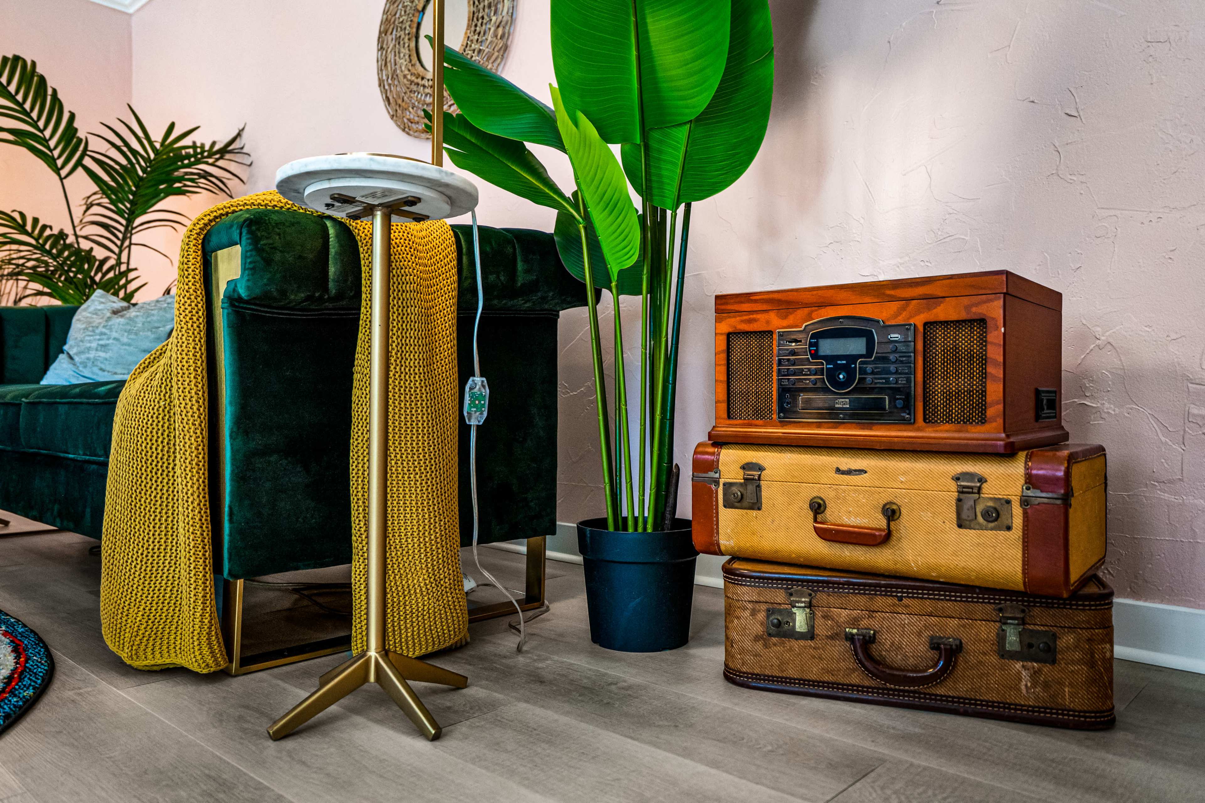 A cozy living room corner featuring a green velvet couch, a decorative potted plant, and a vintage radio atop stacked suitcases.