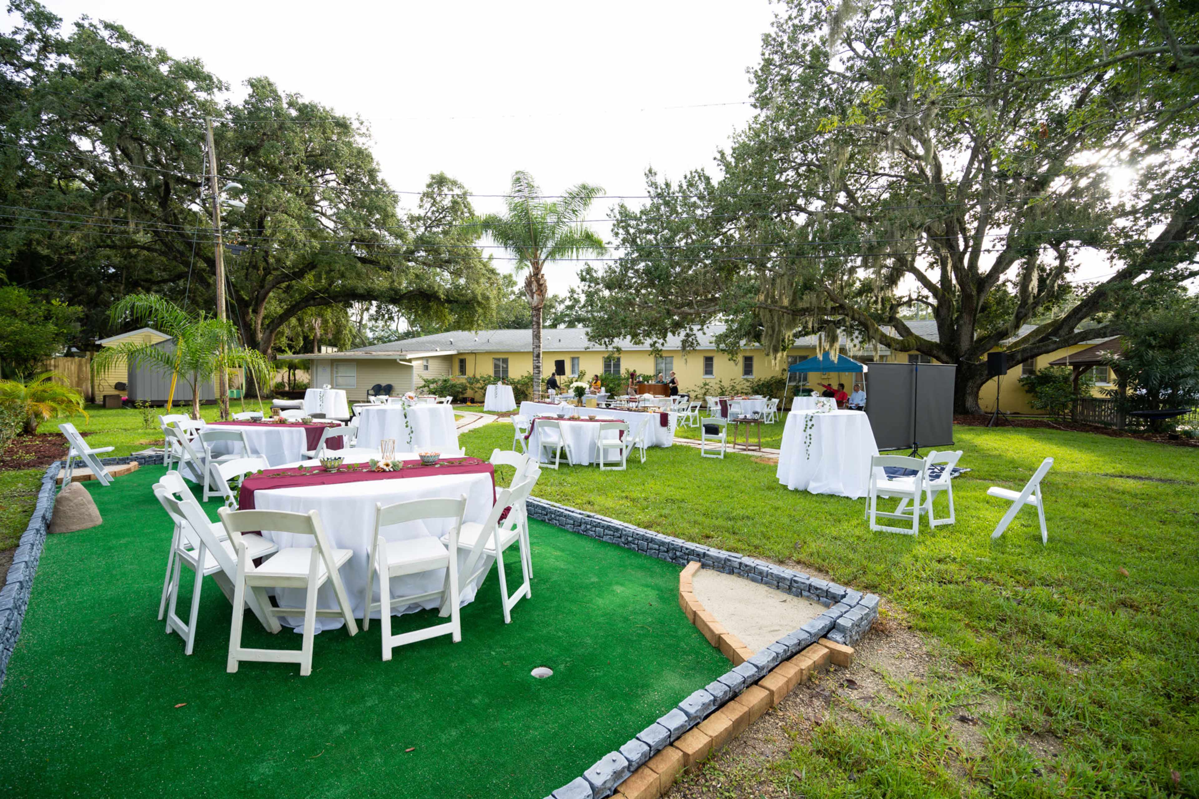 A spacious outdoor area is set up for an event, featuring several round tables covered with white tablecloths and maroon accents placed on green turf.