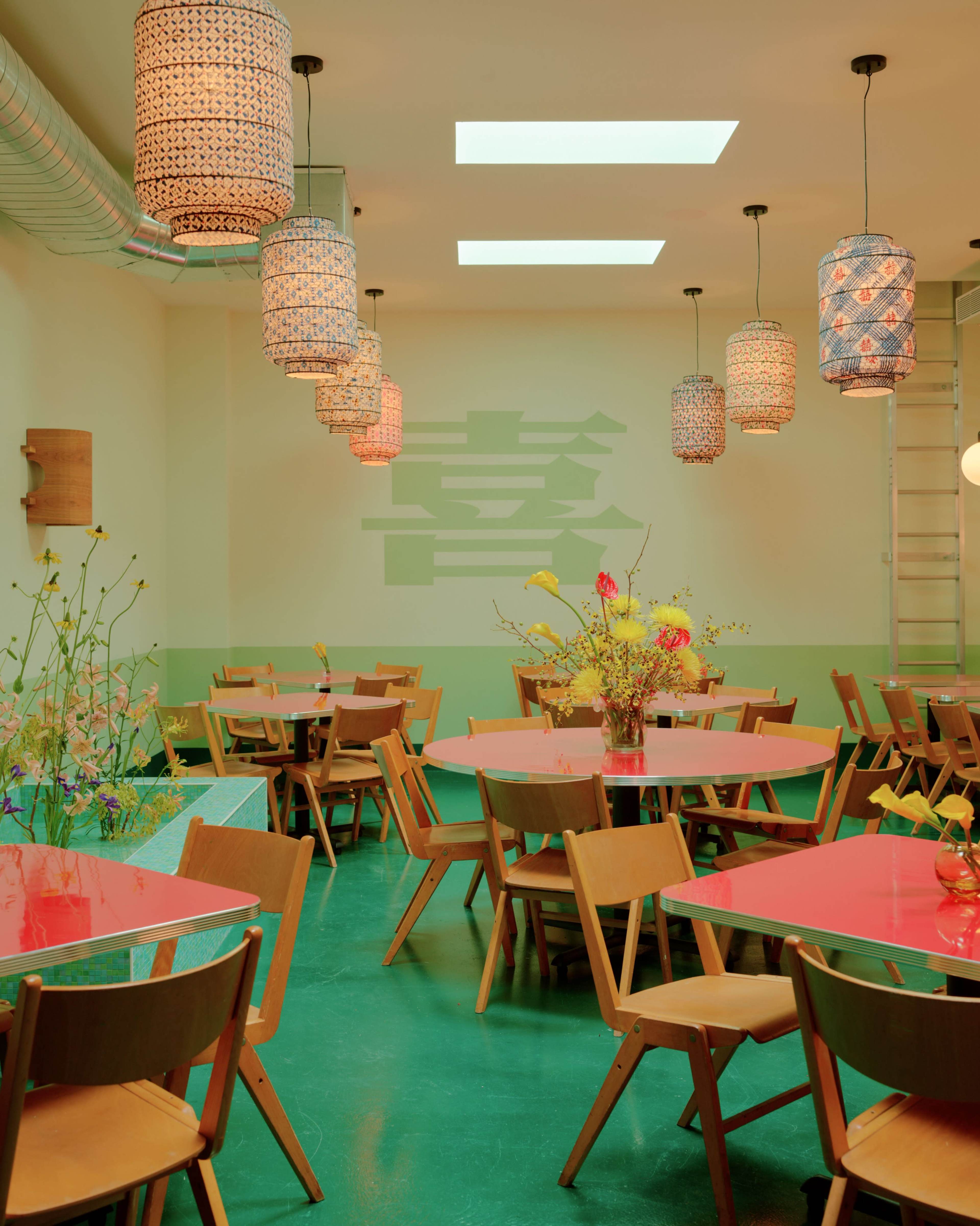 A brightly lit restaurant interior featuring wooden chairs and tables with colorful tabletops, adorned with hanging pendant lights and floral arrangements.