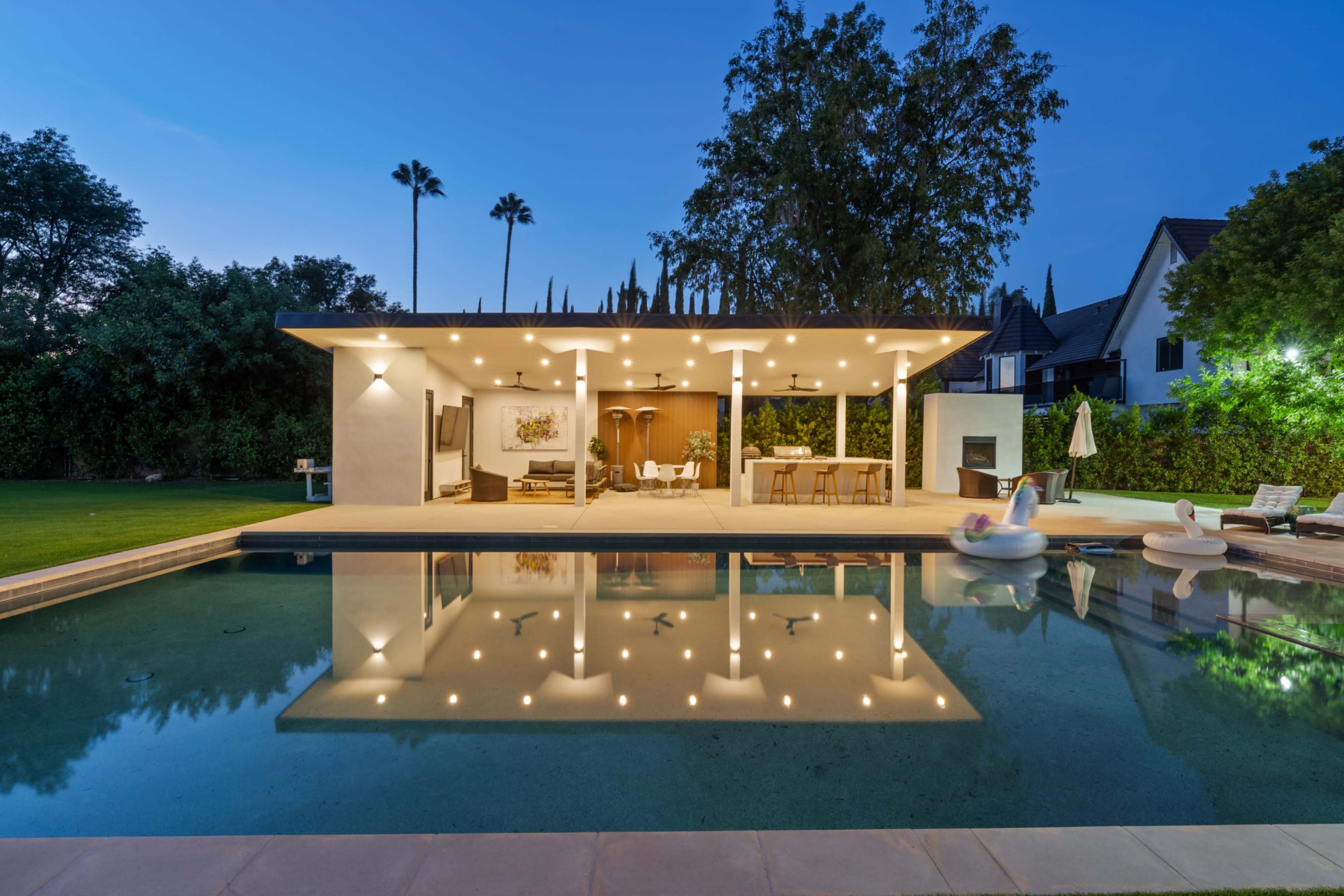 A modern house with a flat roof and large windows is reflected in a still swimming pool, surrounded by a well-kept lawn and palm trees at dusk.