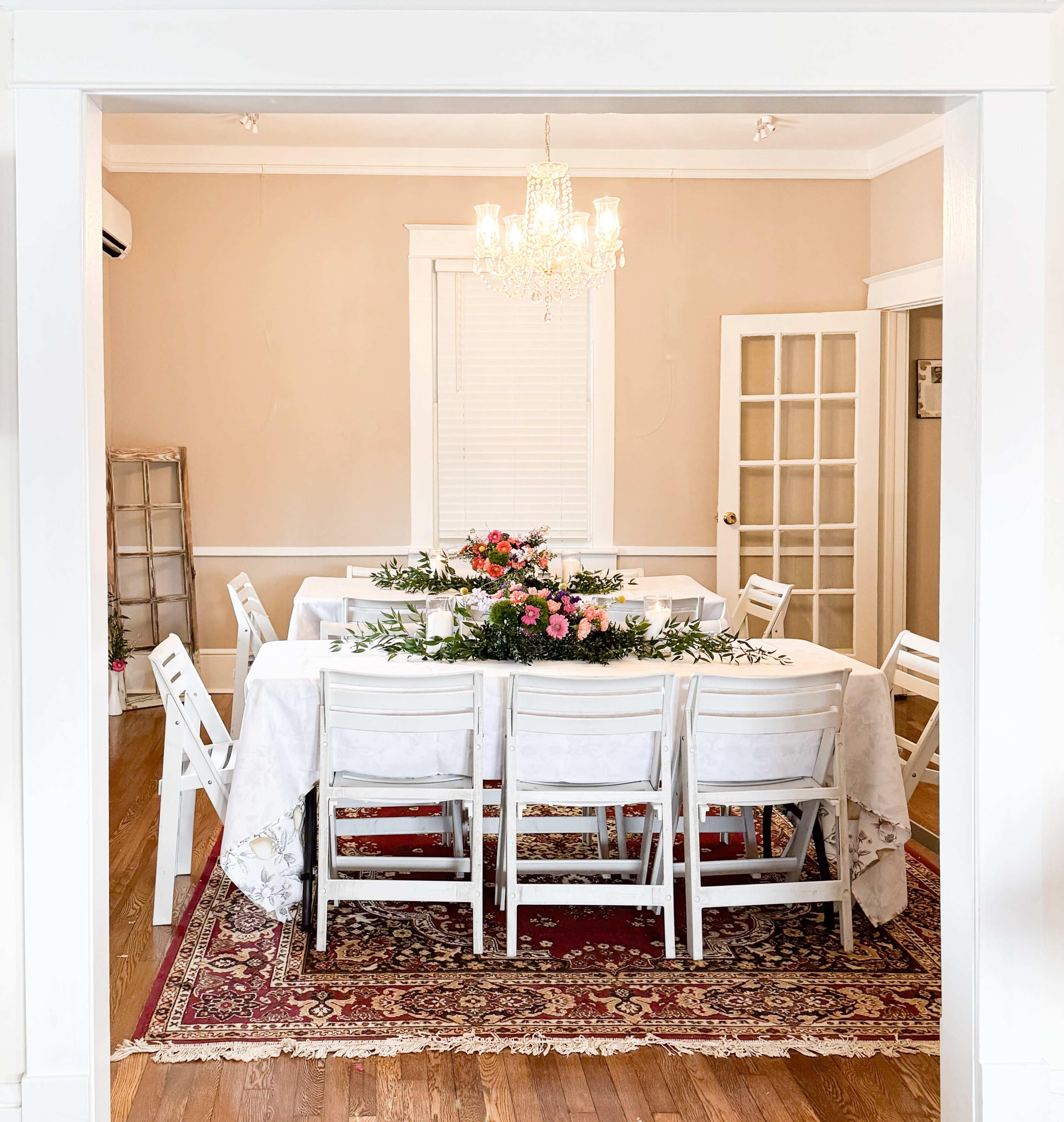 A spacious dining room features a long table set with white tablecloths and decorated with a floral centerpiece, surrounded by white folding chairs on a patterned rug.