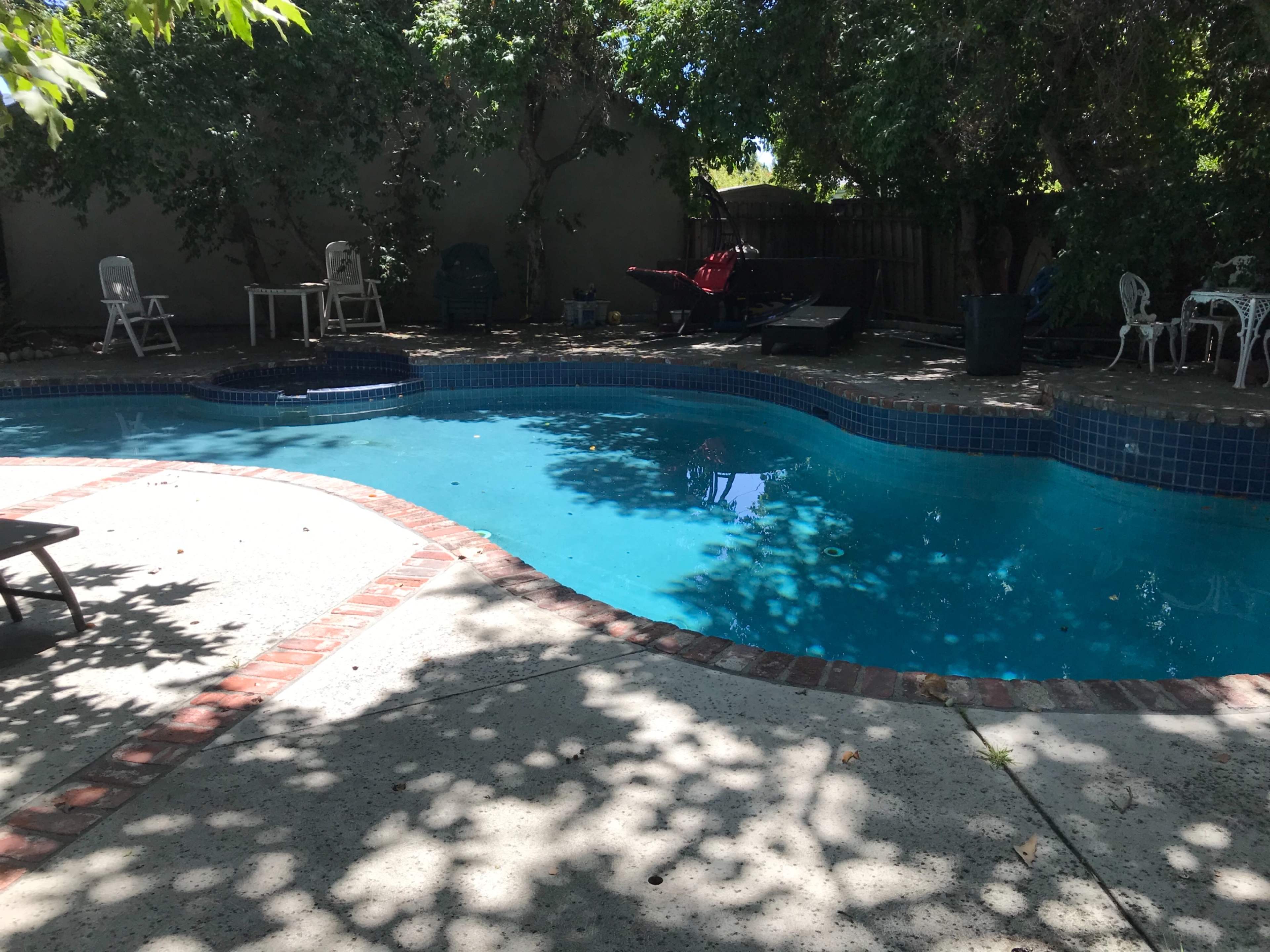 A swimming pool surrounded by trees and patio furniture in a shaded outdoor area.