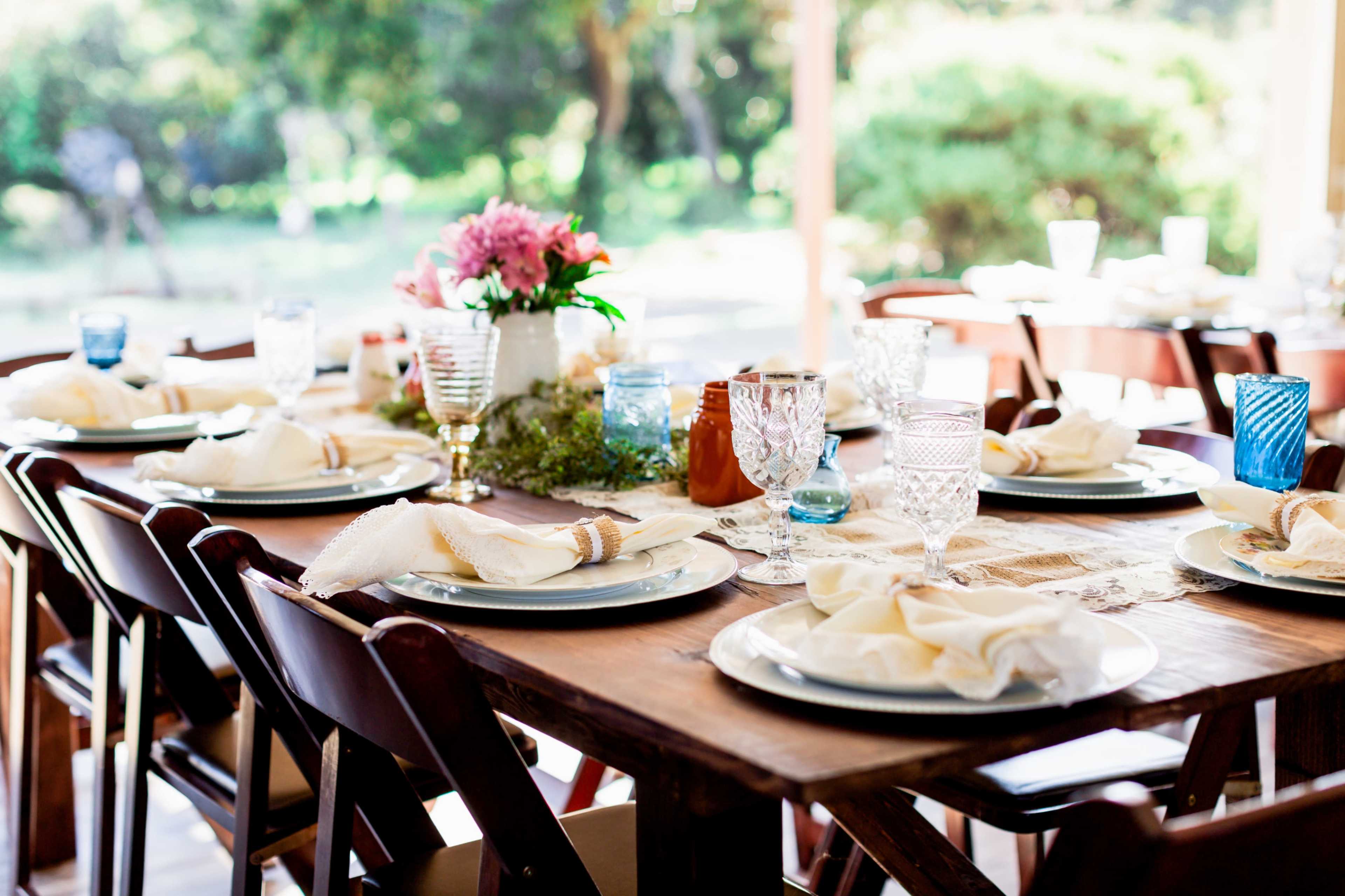 A dining table is set with plates, glasses, and napkins, surrounded by greenery and floral arrangements in a bright outdoor setting.