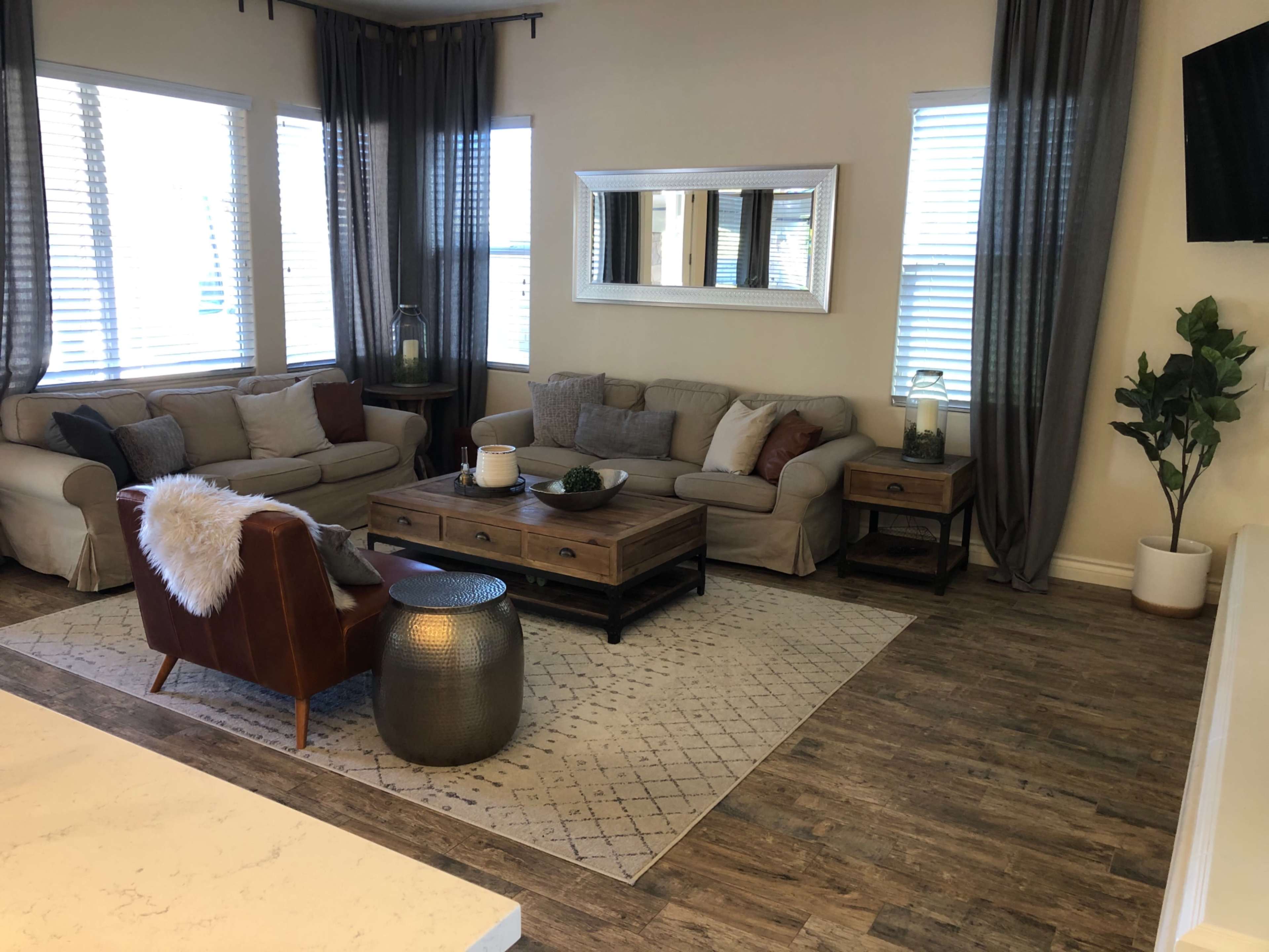 A living room featuring a sectional sofa, a coffee table, a plant, and large windows with sheer curtains.