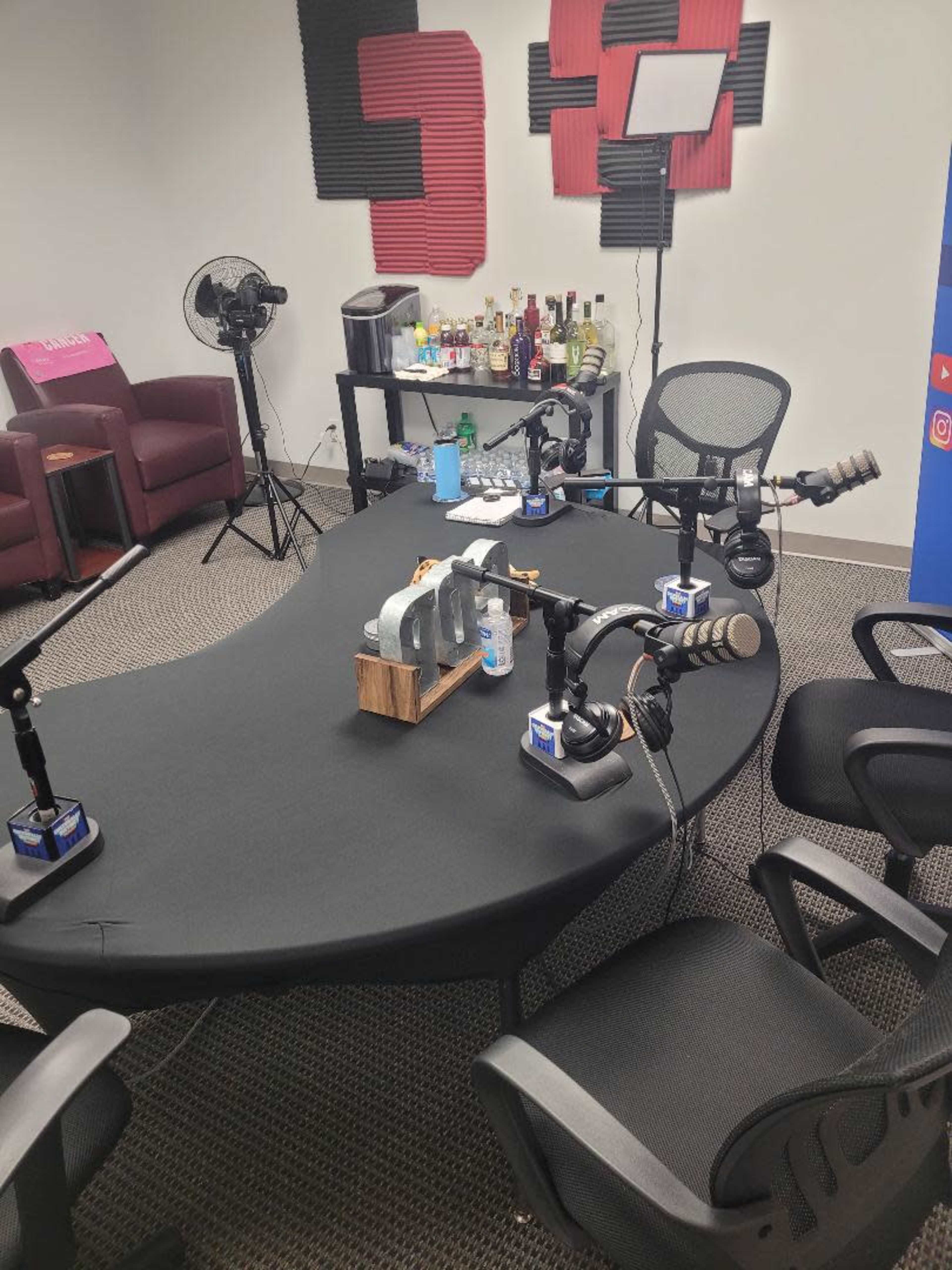 A podcast recording setup is shown, featuring microphones on arm stands, chairs, and a table with various drink bottles in a room decorated with soundproofing panels.