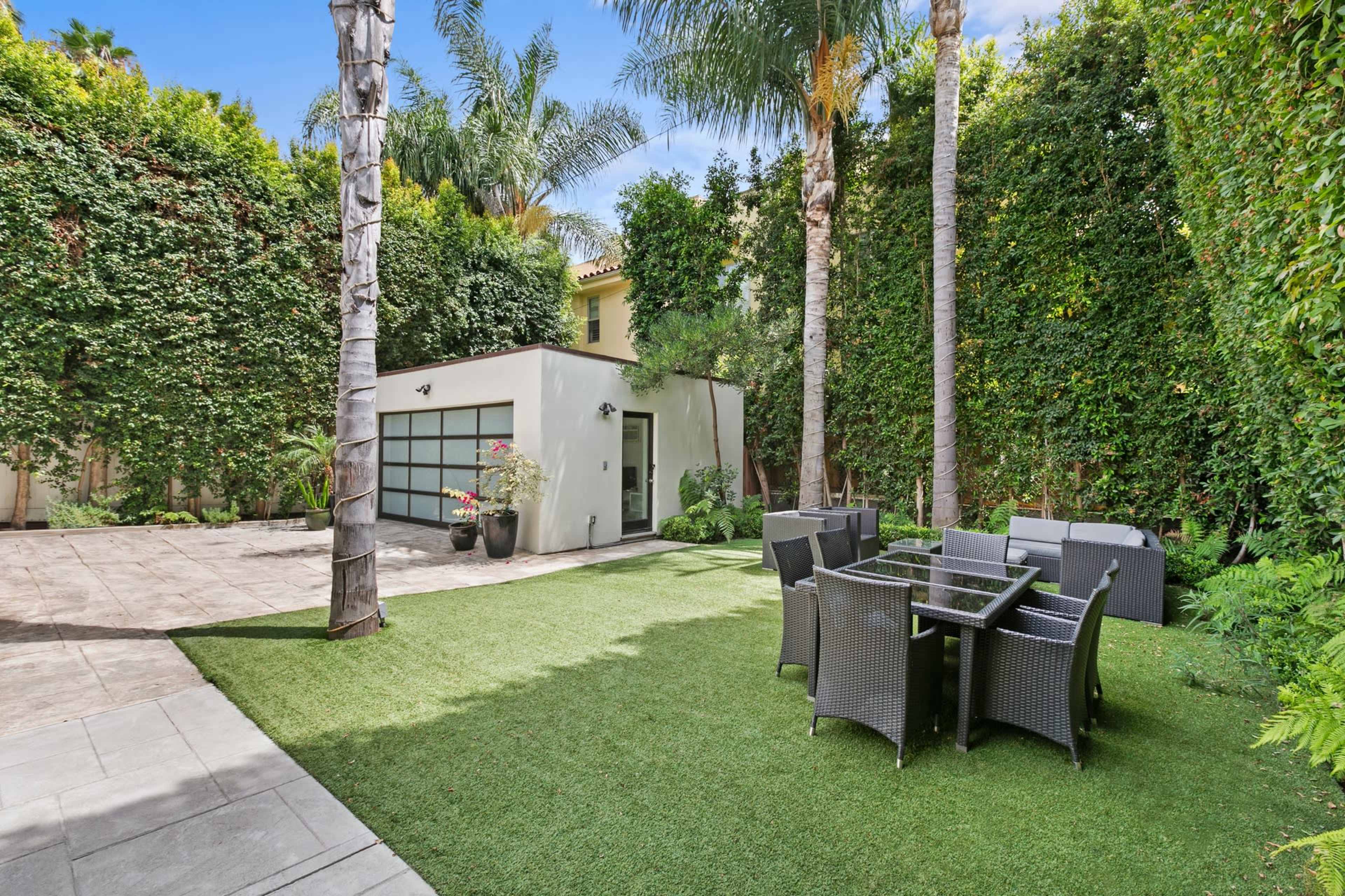 A landscaped outdoor area features a small building, a grassy lawn, and a dining set surrounded by tall palm trees and vibrant greenery.