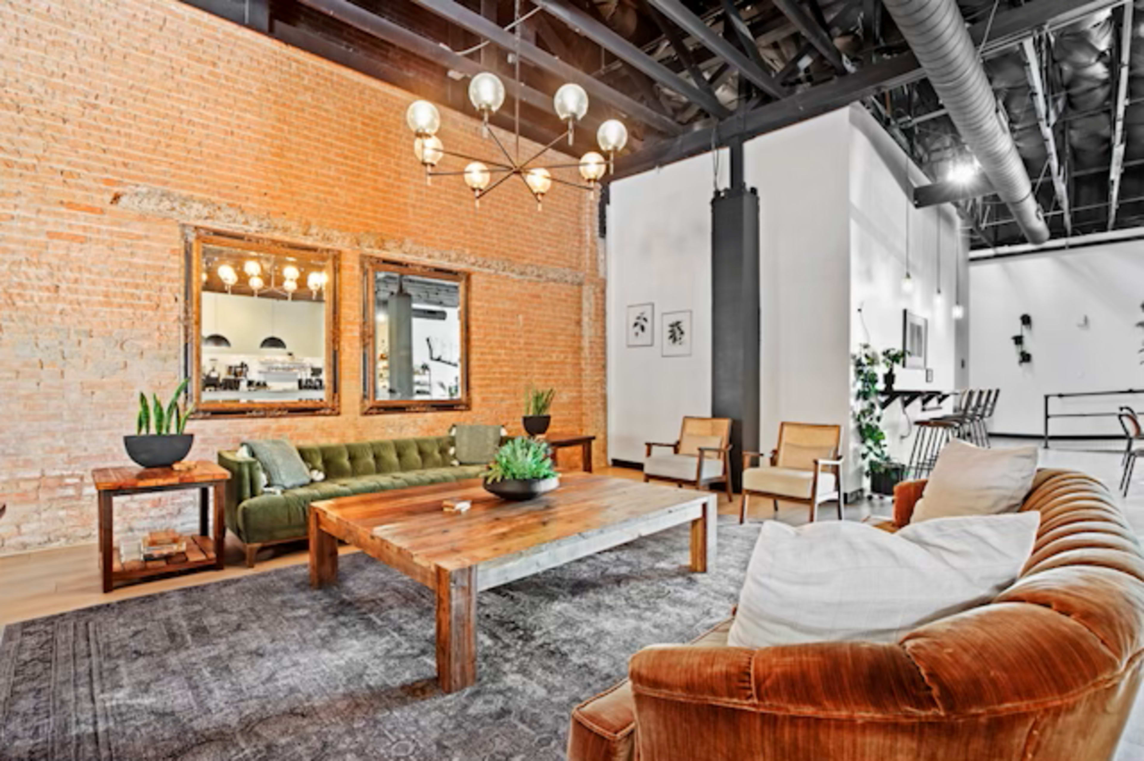 The image shows a modern living space with exposed brick walls, a wooden coffee table, a green sofa, and two armchairs, all arranged around a rug.