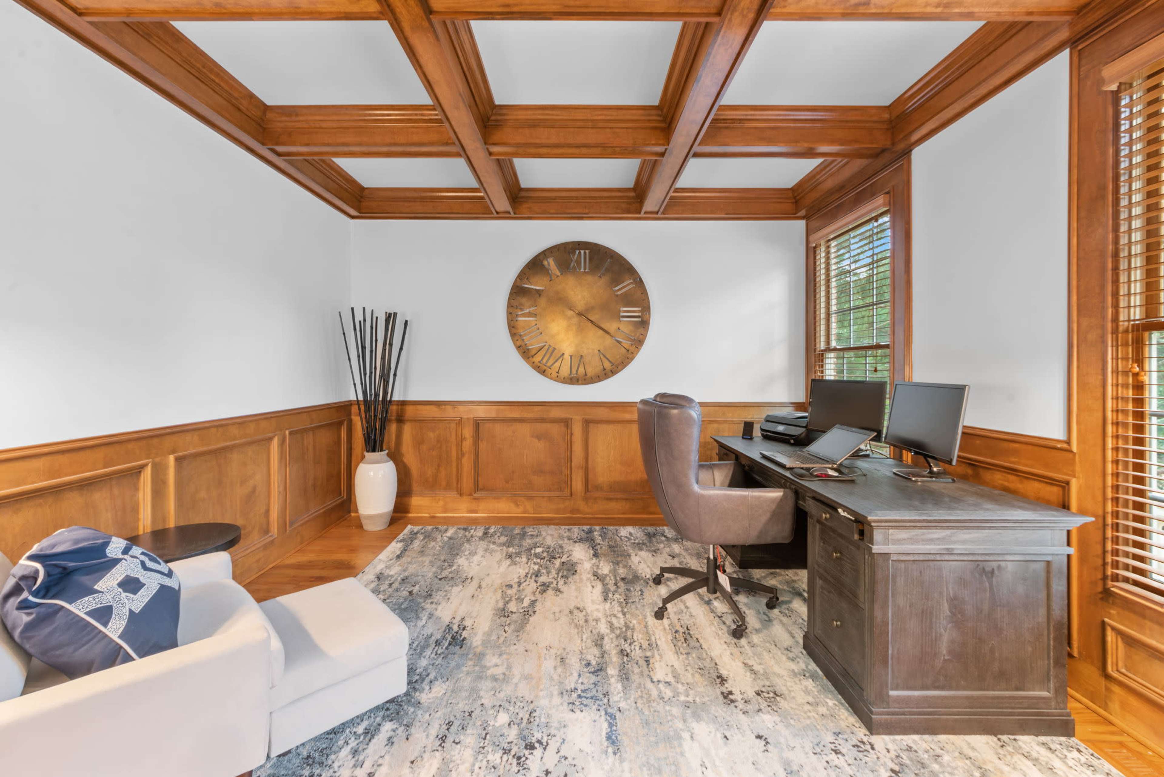 A home office with a wooden coffered ceiling, a large wall clock, two computer monitors on a desk, and a light-colored sofa.