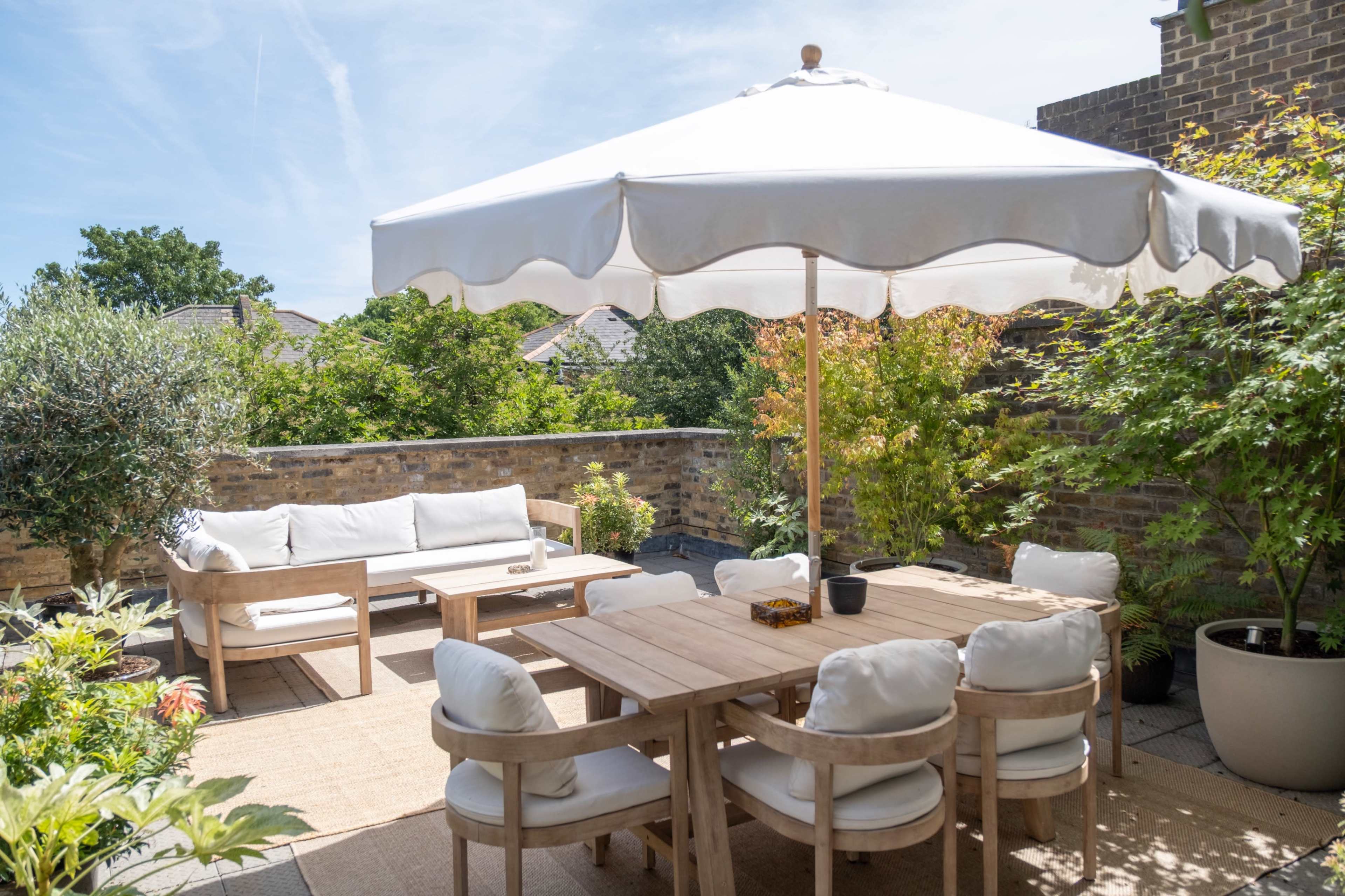 A wooden dining table and chairs are set under a large white umbrella in a sunlit outdoor patio surrounded by greenery.