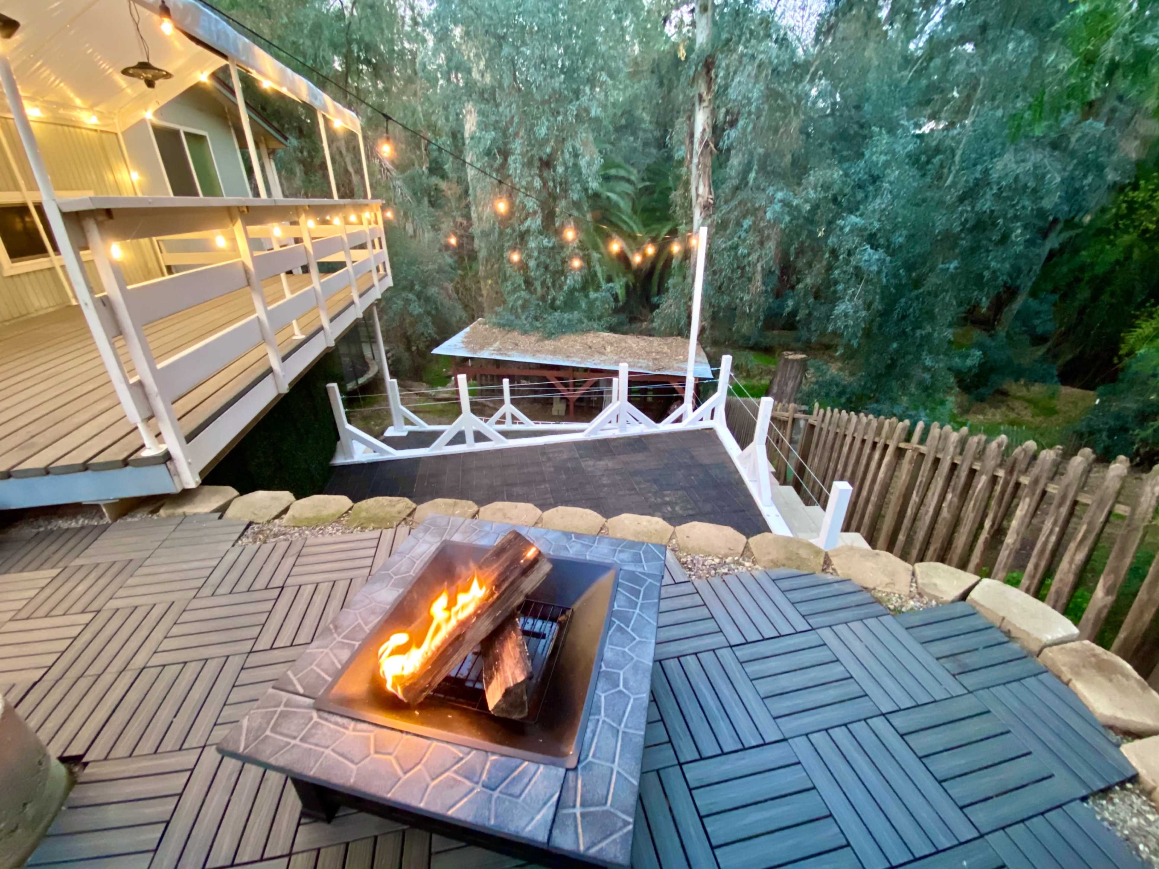 A fire pit with burning logs sits in the foreground, overlooking a patio and a deck illuminated by string lights among trees.