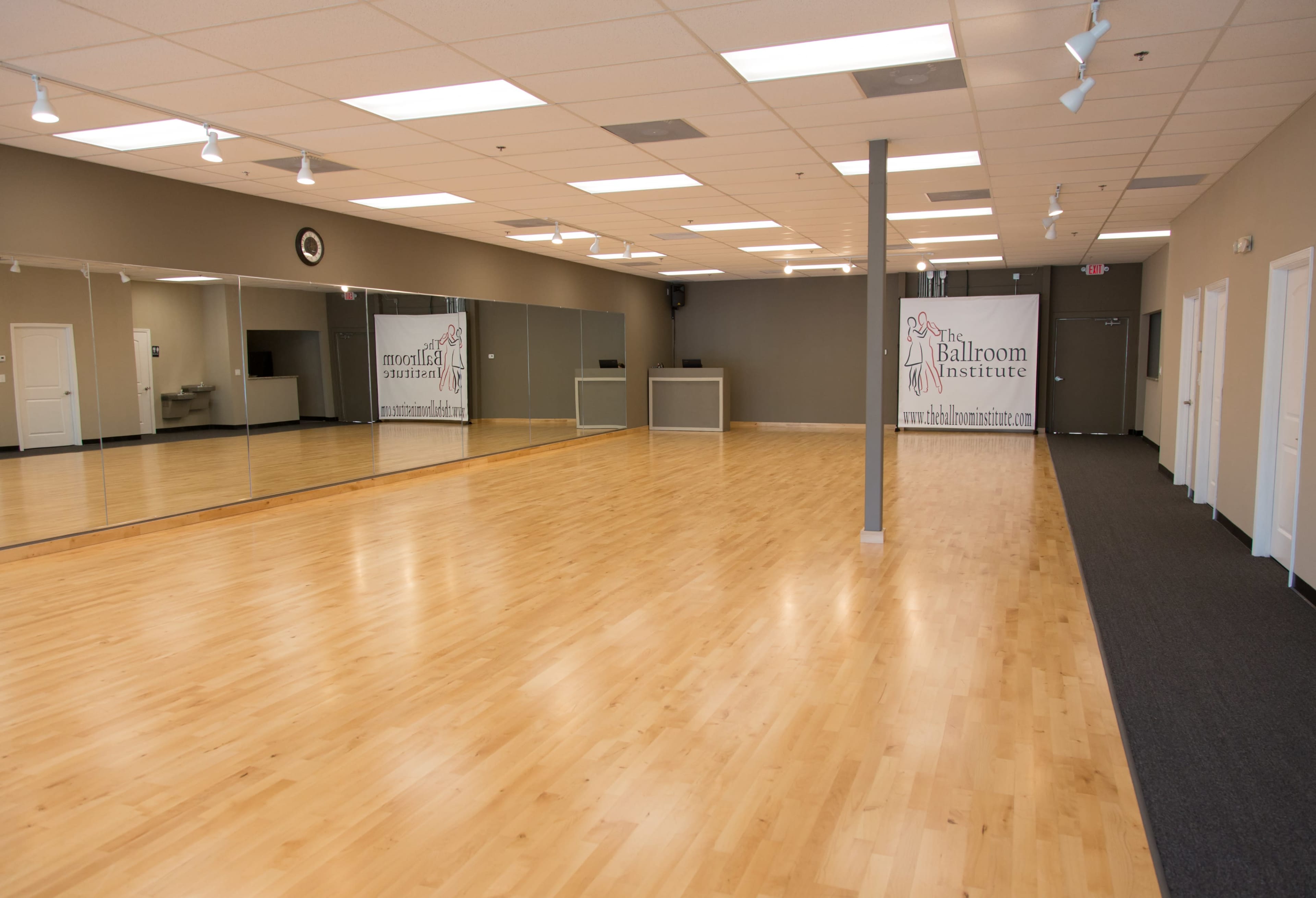 The image shows an empty dance studio with wooden flooring, large mirrors on one wall, and a banner for a ballroom institute.