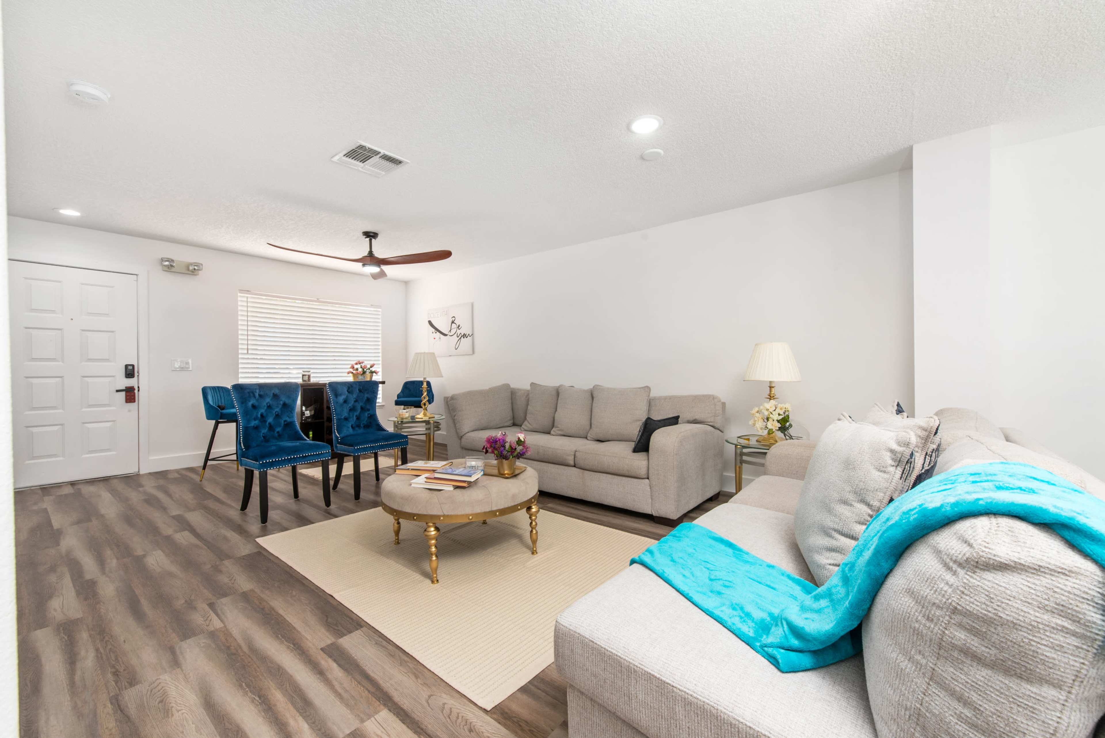 A living room features two sofas, a coffee table, and a dining area with blue chairs, illuminated by natural light.