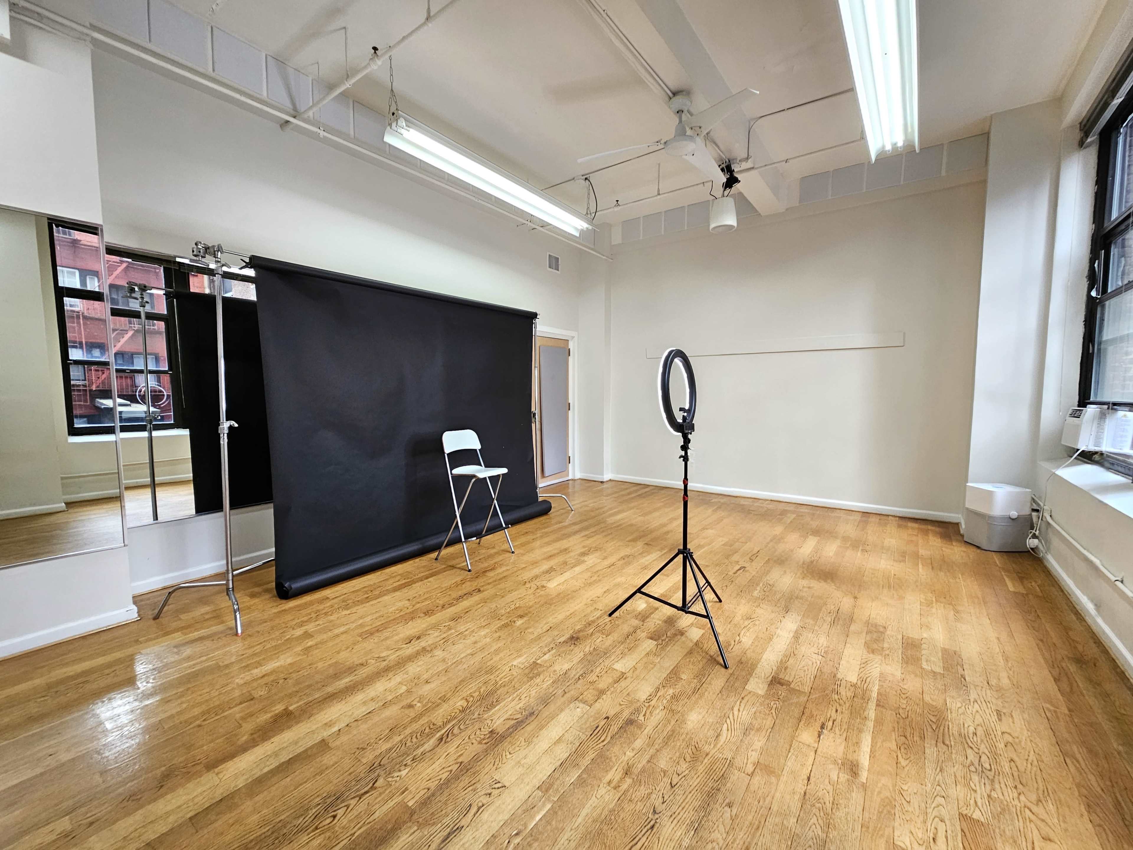 The image shows a spacious, empty room with wooden flooring, a black backdrop on a stand, a folding chair, and a ring light set up near a large window.