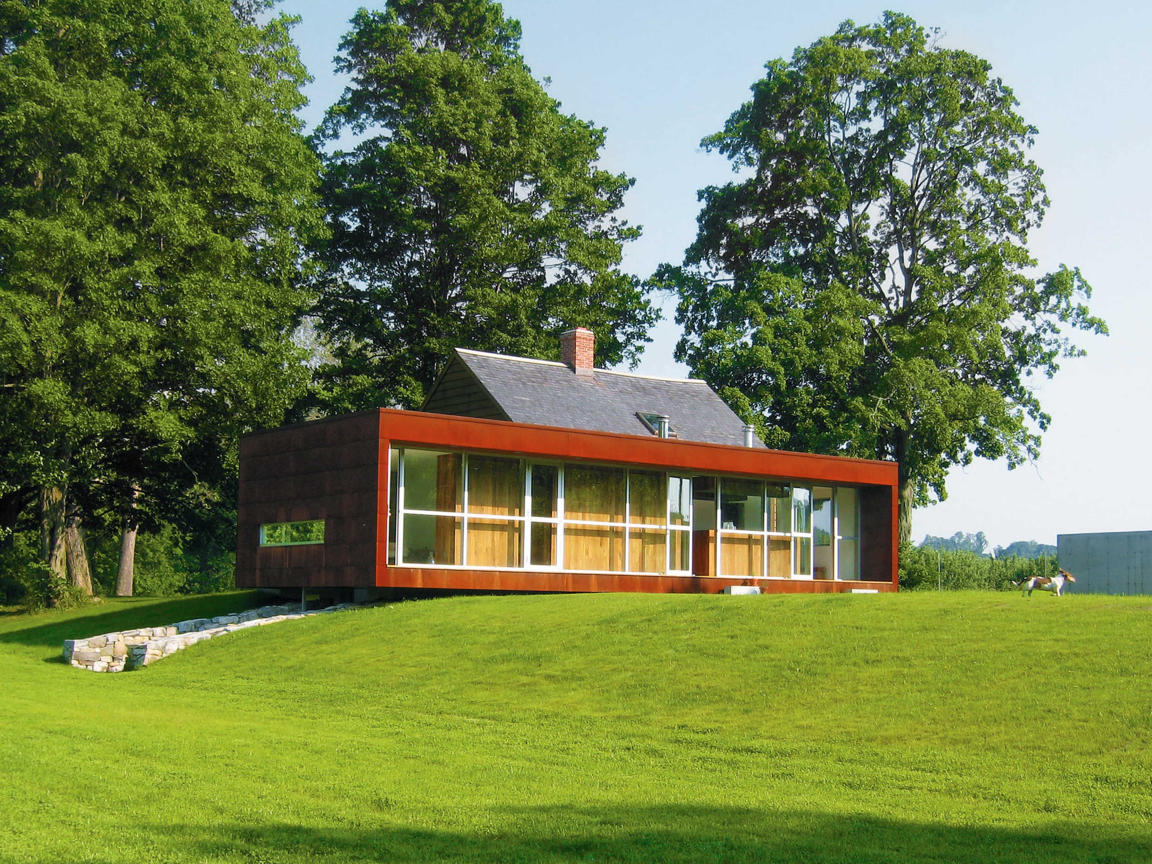 A modern house with a combination of wooden and glass elements sits on a grassy slope, surrounded by large trees.