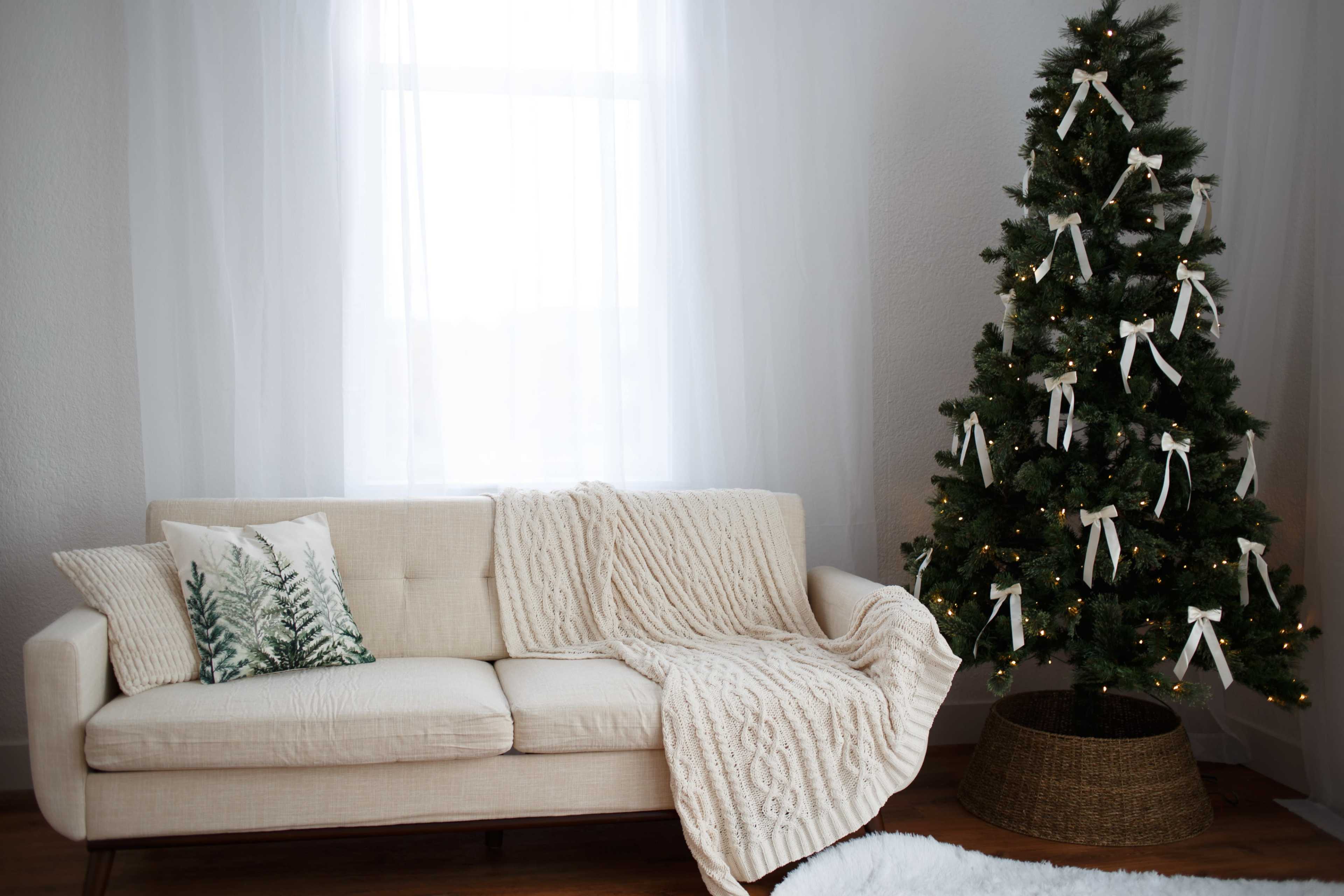 A beige couch with a knitted throw and a decorative pillow is positioned next to a Christmas tree adorned with white ribbons and lights, set against a window with sheer curtains.