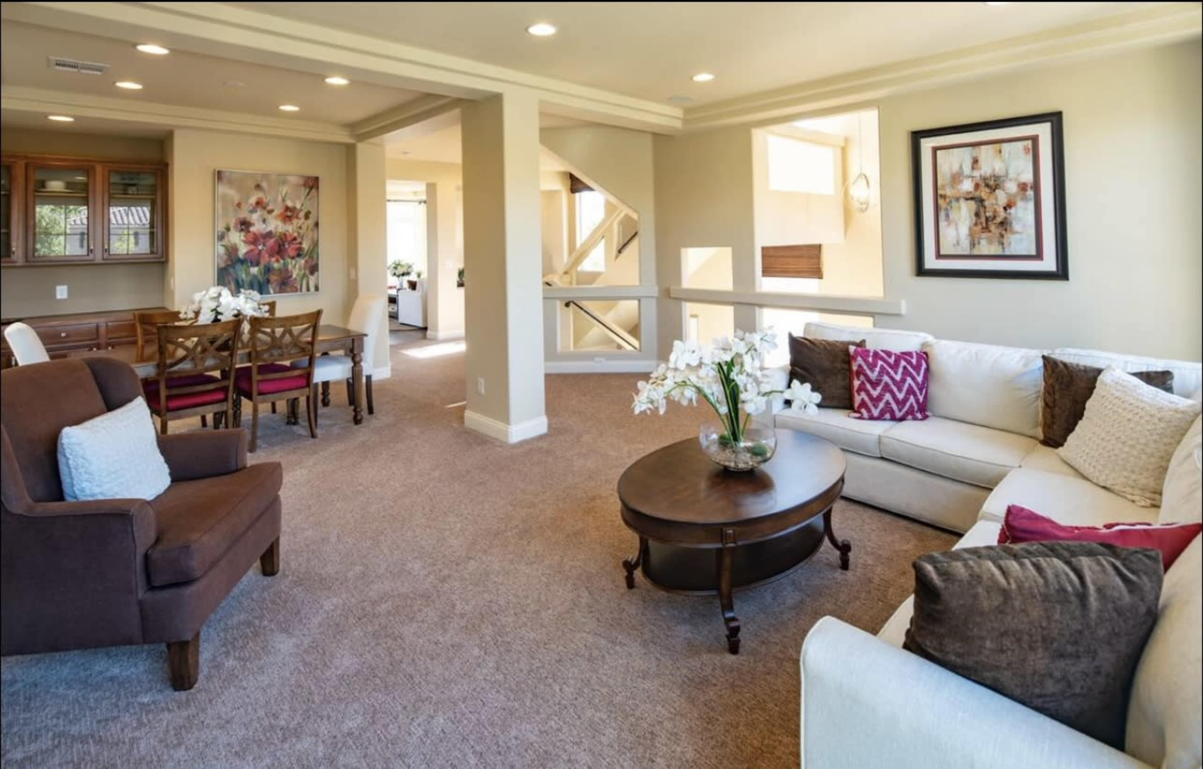 The image shows a bright, open living space featuring a white sectional sofa, a round coffee table with flowers, and a dining area with wooden chairs.