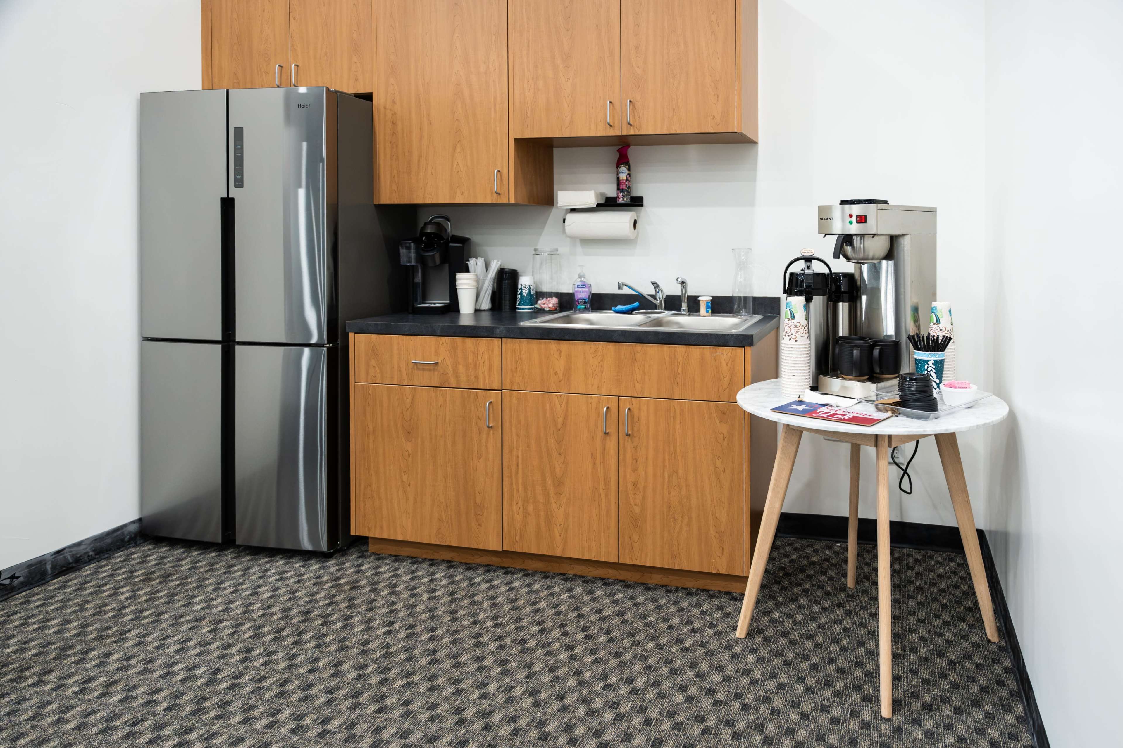 The image shows a kitchenette area with a stainless steel refrigerator, wooden cabinets, a sink, and a coffee maker on a small table.