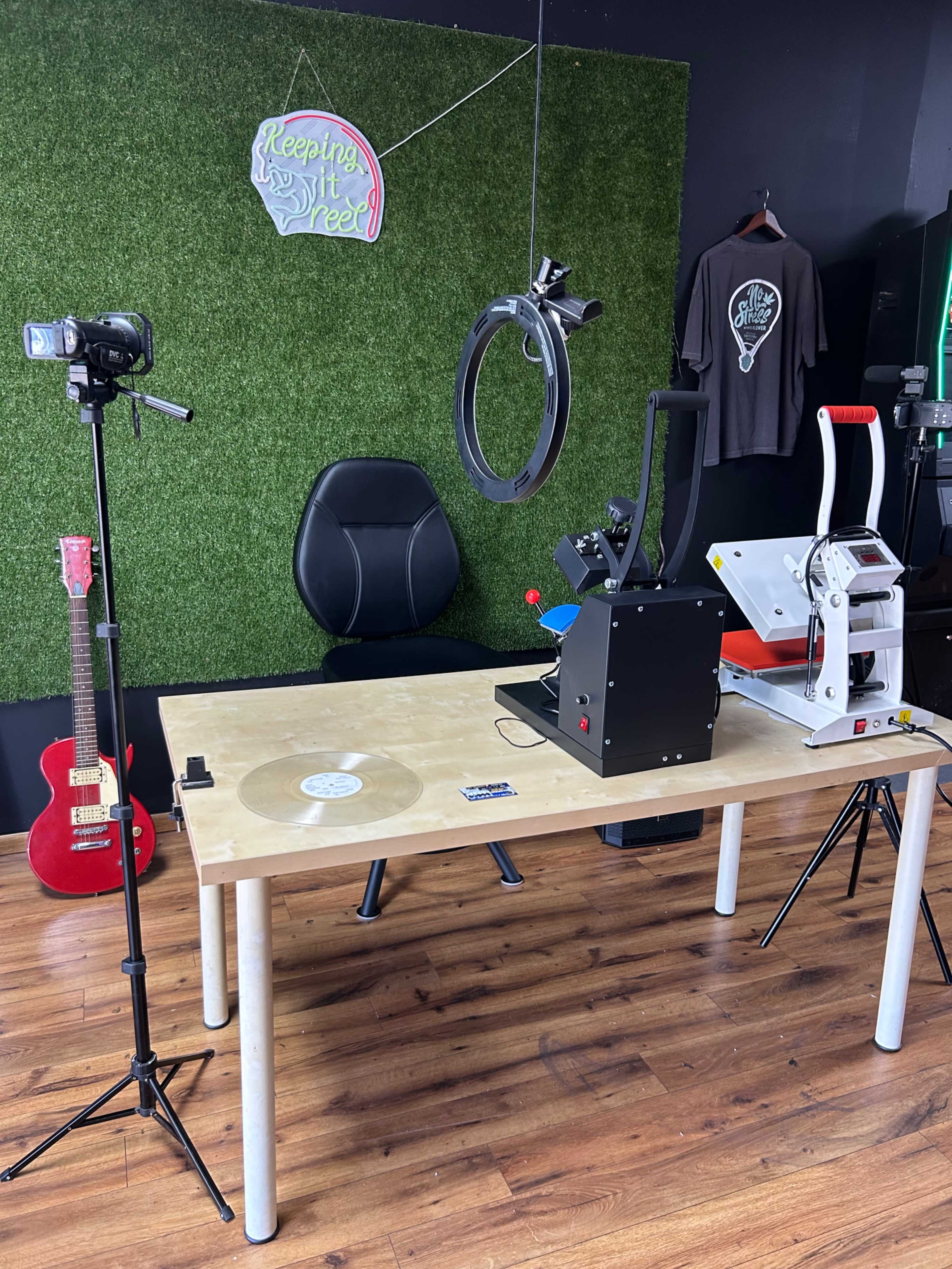 The image shows a workspace with a wooden table equipped with a vinyl press, a ring light, two cameras on tripods, and a black chair against a backdrop of green turf.