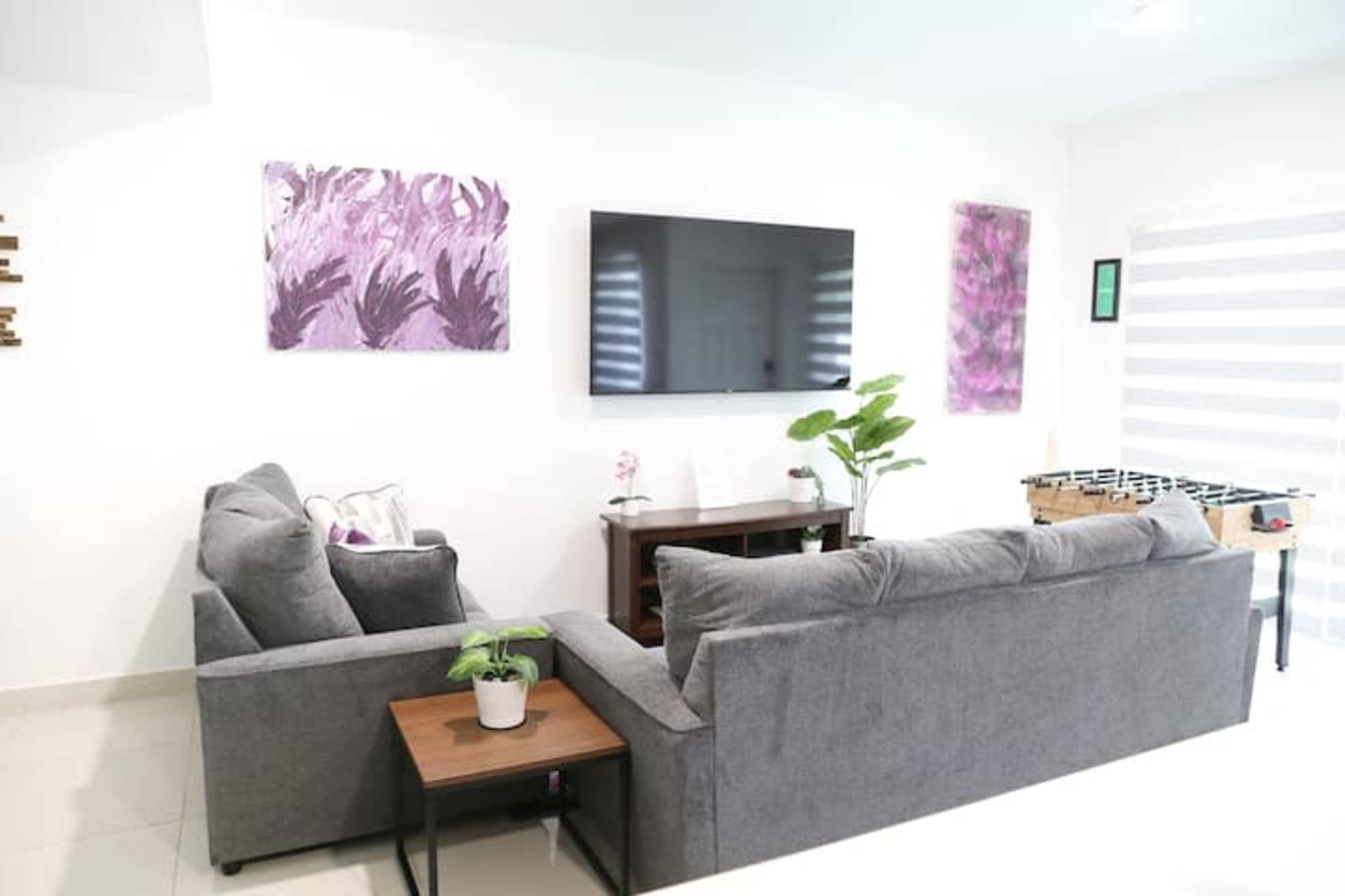 A modern living room features two gray sofas, a wall-mounted TV, and abstract artwork in purple, with a small table and a potted plant.
