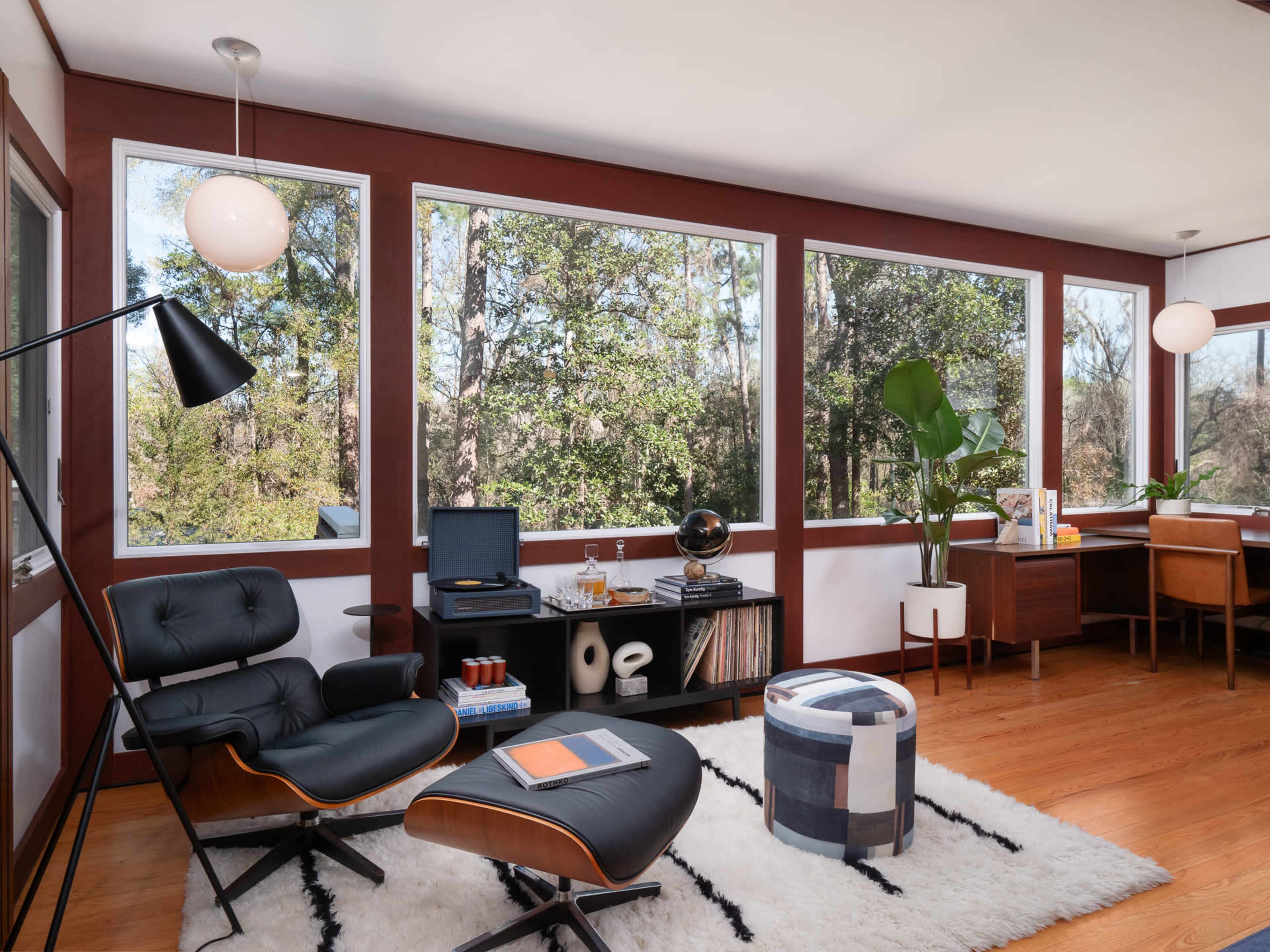 A modern living room features a black lounge chair and ottoman, a wooden desk, and large windows overlooking a forested area.