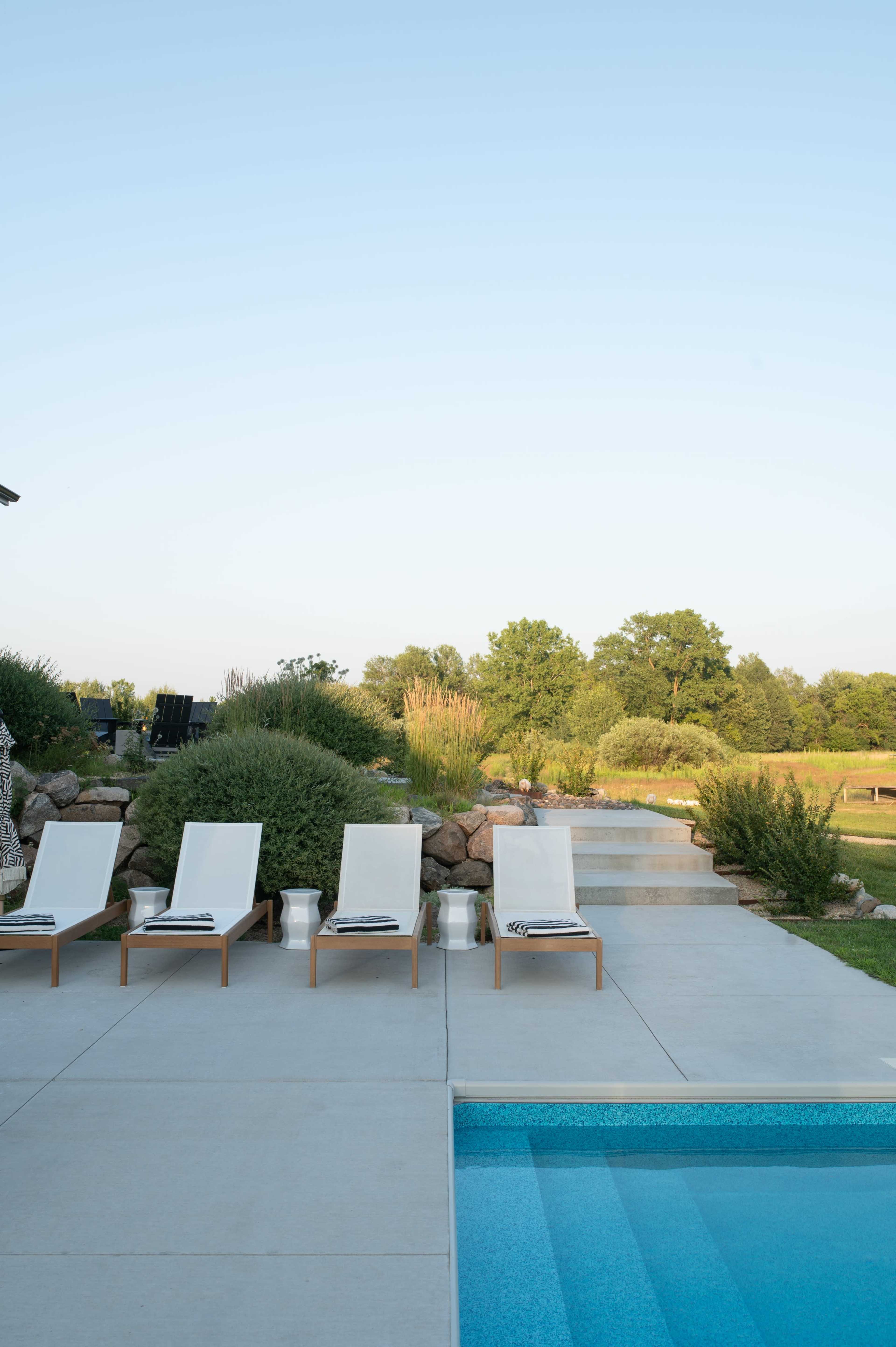 The image shows a tranquil outdoor setting featuring four lounge chairs by a swimming pool, surrounded by landscaped greenery and rocks.