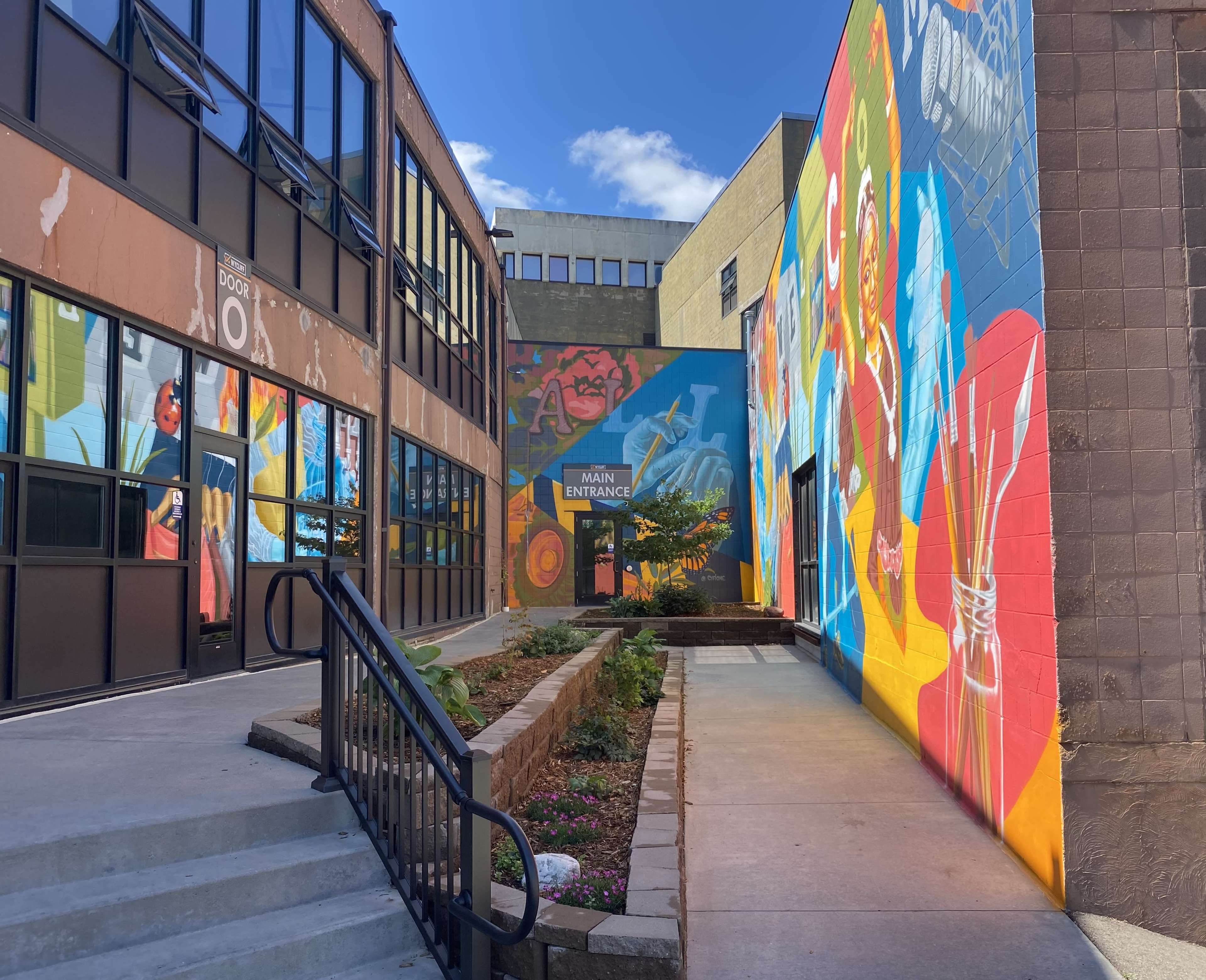 A narrow walkway leading to a brightly painted mural wall, flanked by buildings with large windows and a small garden area.