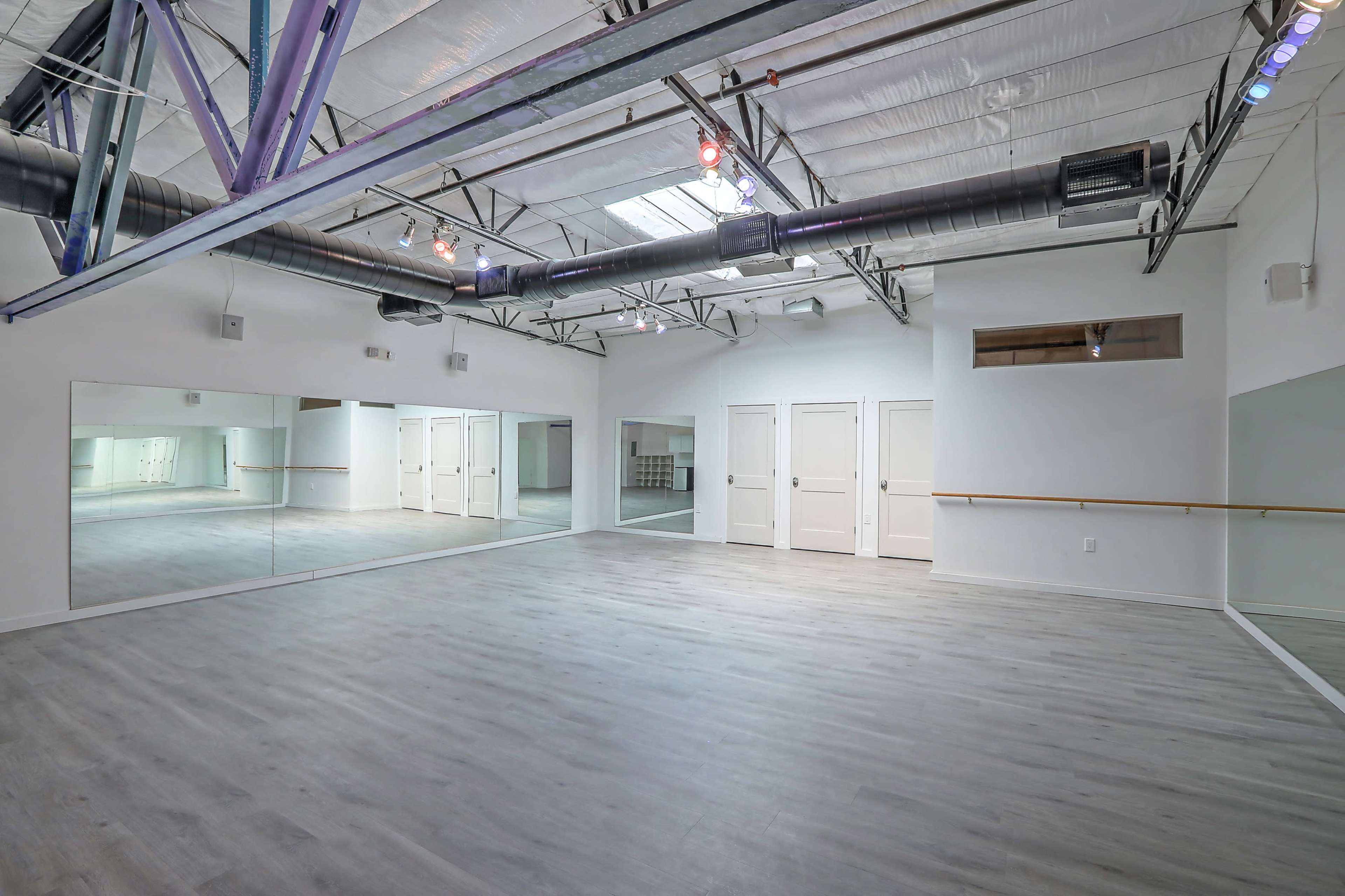 A spacious dance studio features hardwood flooring, mirrors on one wall, and white walls with a ceiling exposed to metal beams.