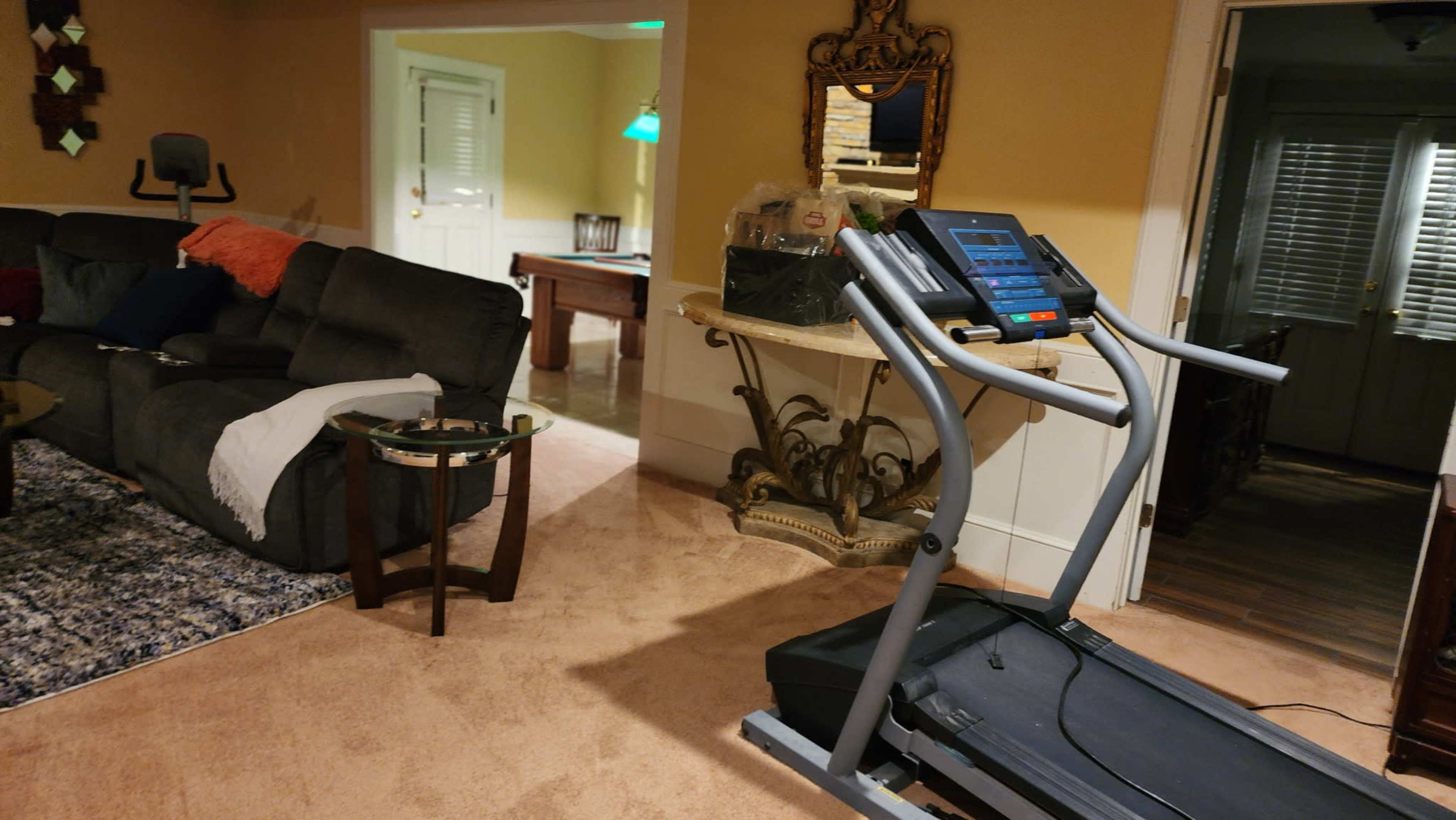 A treadmill is positioned in a living area adjacent to a sectional sofa and a table near an open door leading to another room.
