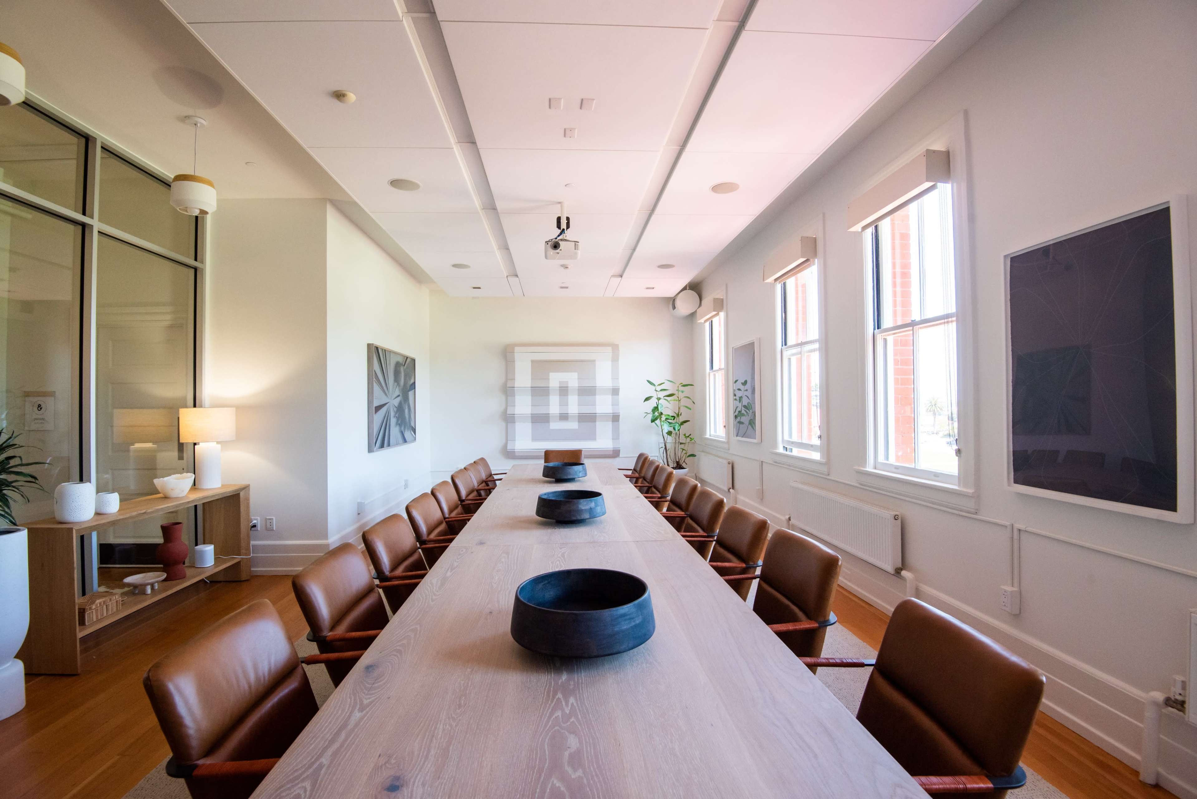 A long wooden conference table surrounded by leather chairs is set in a brightly lit meeting room with large windows and modern decor.