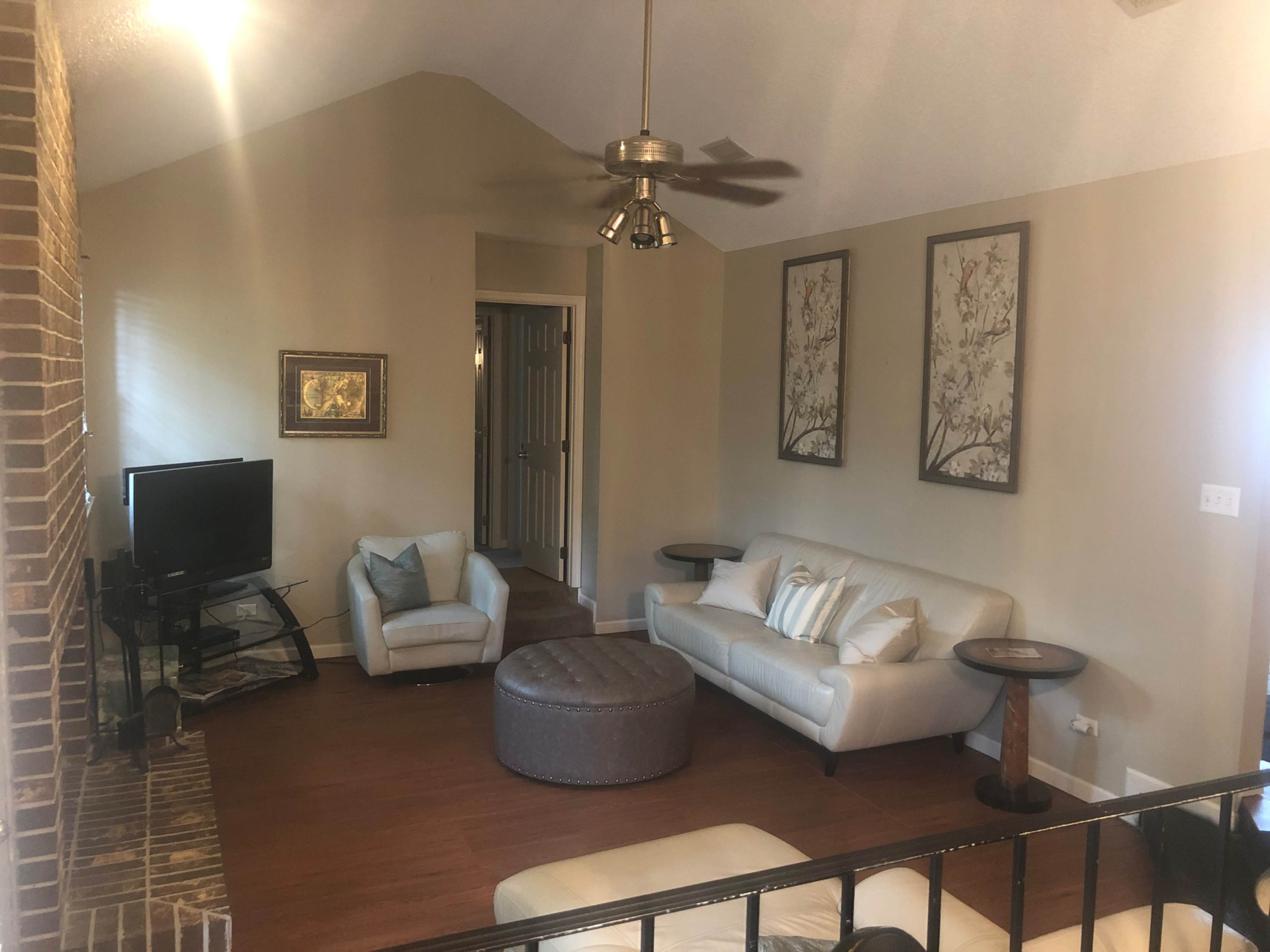 The image shows a living room with a TV, a light-colored sofa set, a round ottoman, and two decorative wall art pieces.