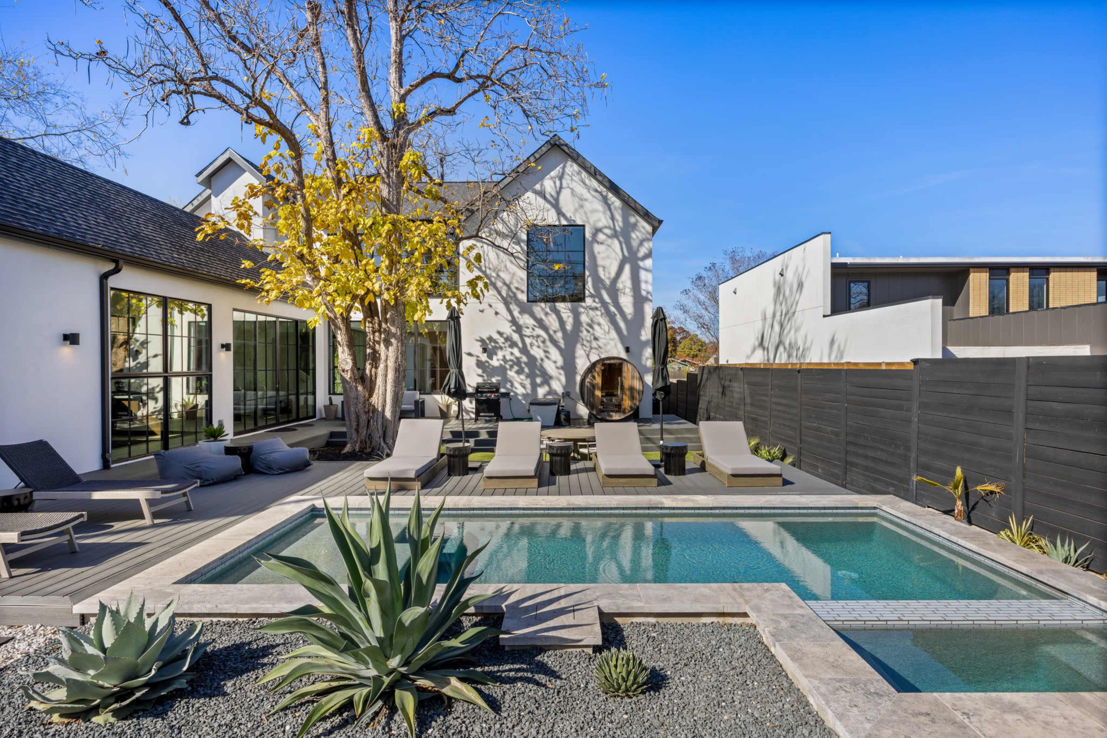 A modern backyard features a swimming pool surrounded by lounge chairs, rocks, and plants, with a house in the background.