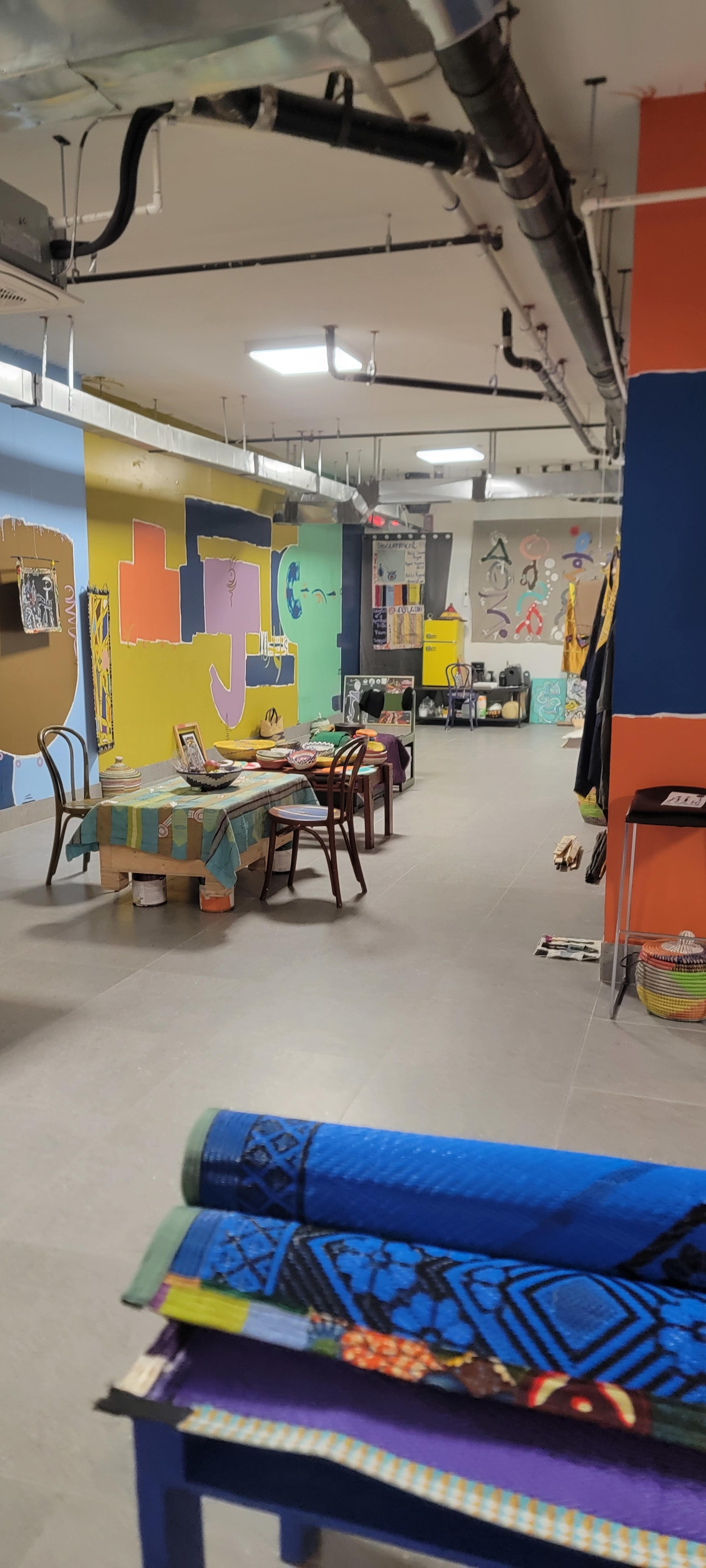 The image shows a spacious room with colorful walls and various tables, chairs, and art supplies scattered throughout.