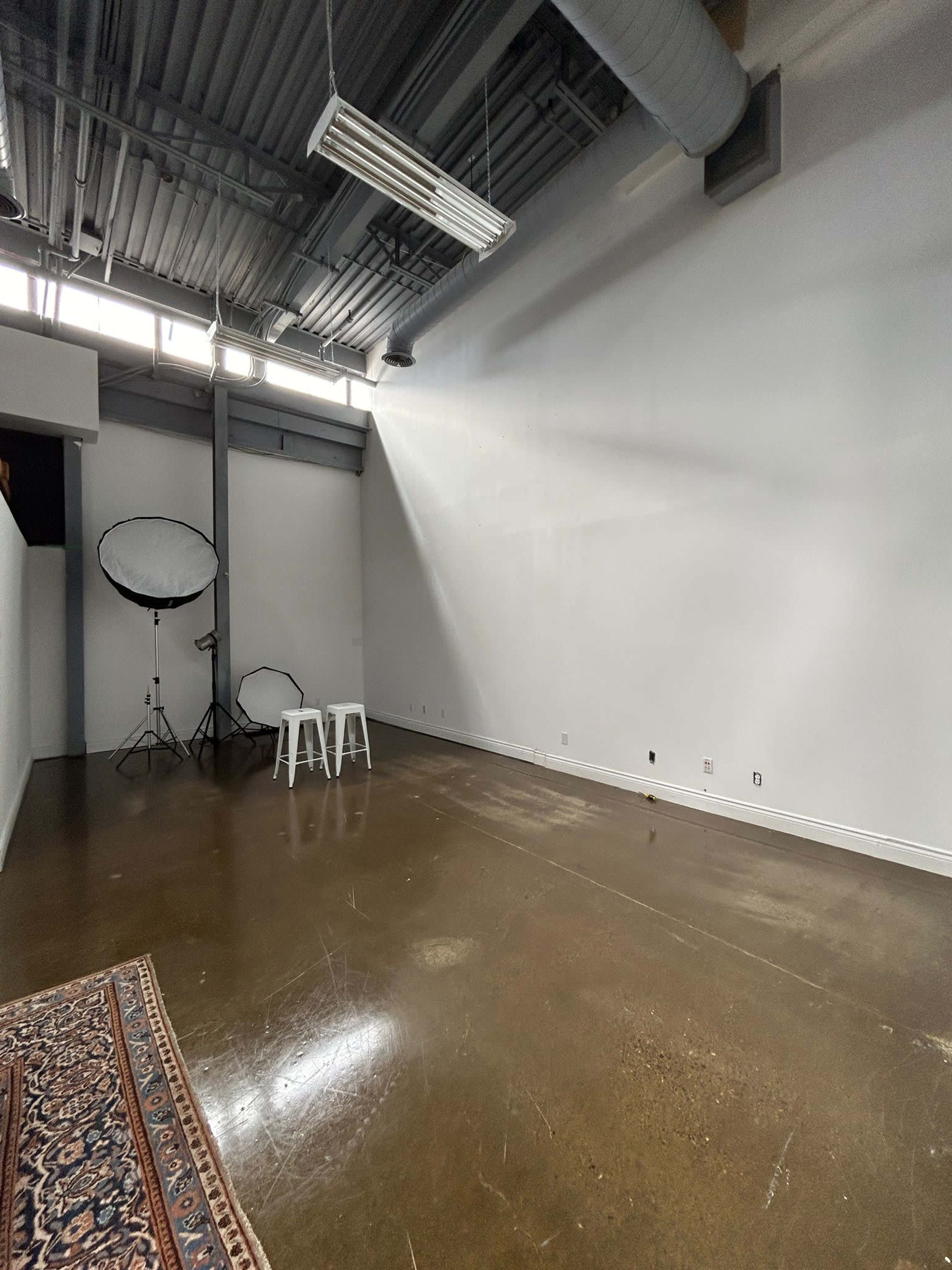 The image shows a spacious, empty room with a polished concrete floor, a light-colored wall, two white stools, and photography equipment in the background.