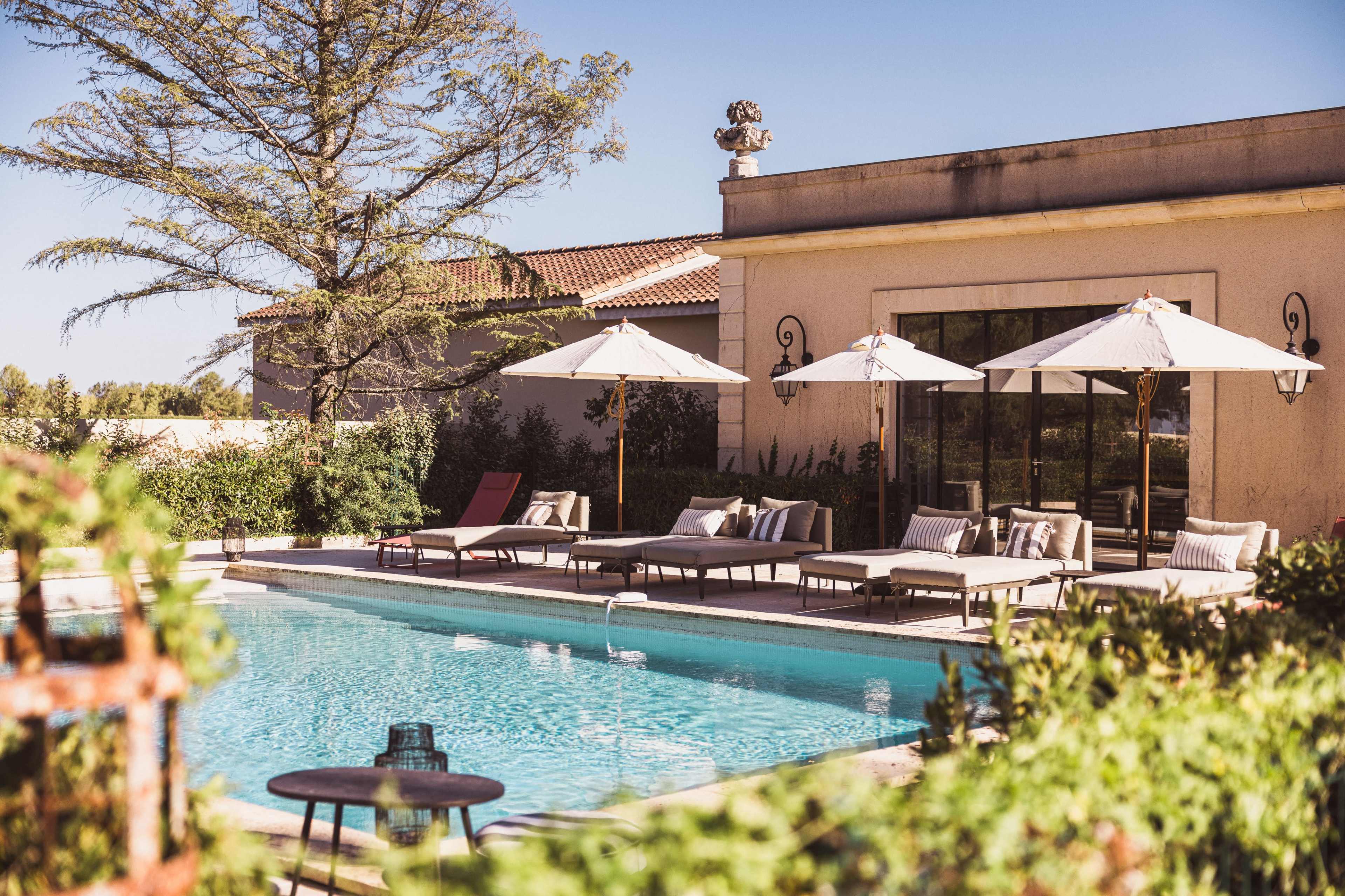 The image shows a residential pool area with lounge chairs and umbrellas surrounding a clear blue pool, framed by greenery and a sunny sky.