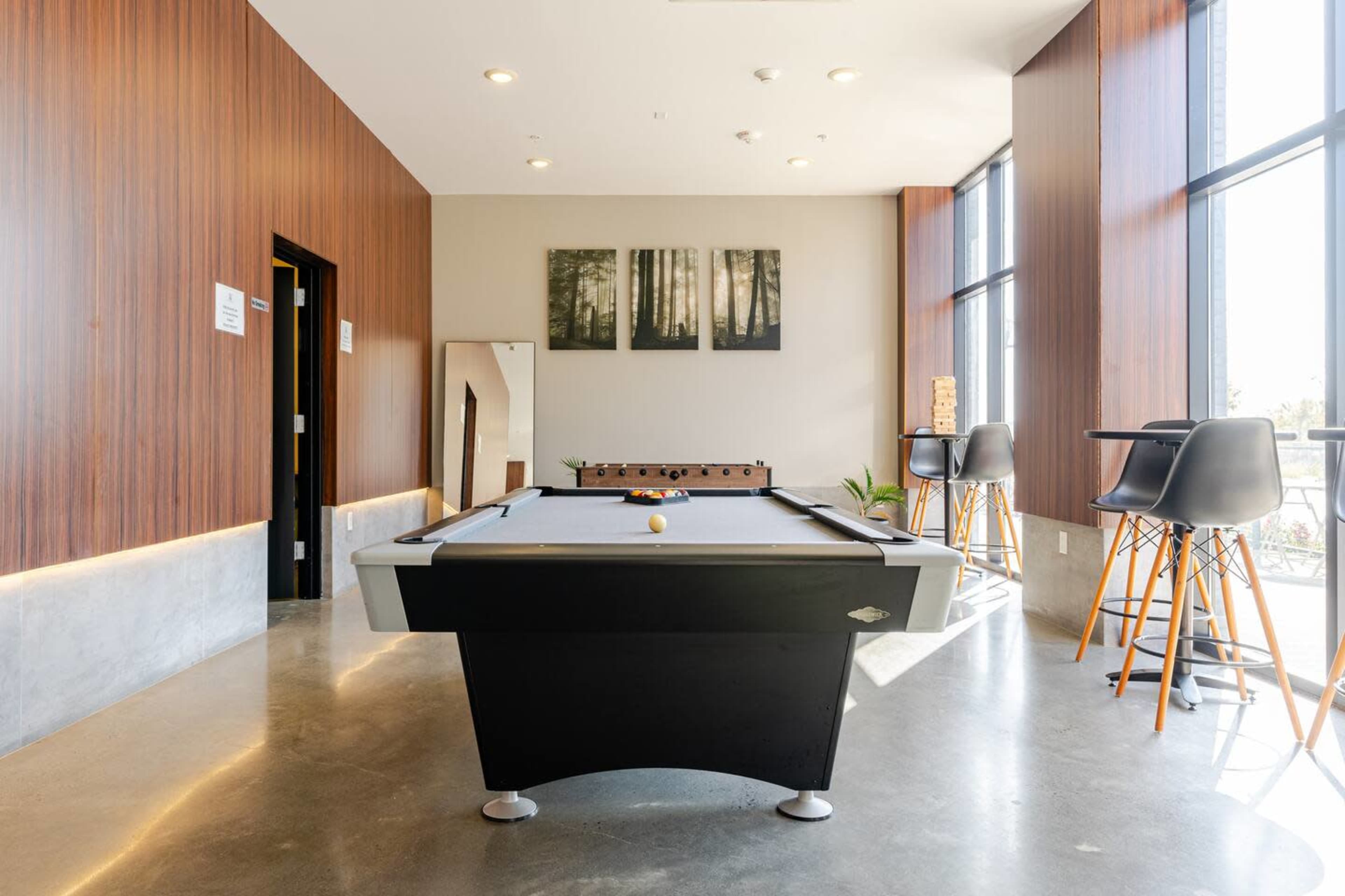 The image features a pool table set in a modern room with wooden paneling, large windows, and a few barstools.