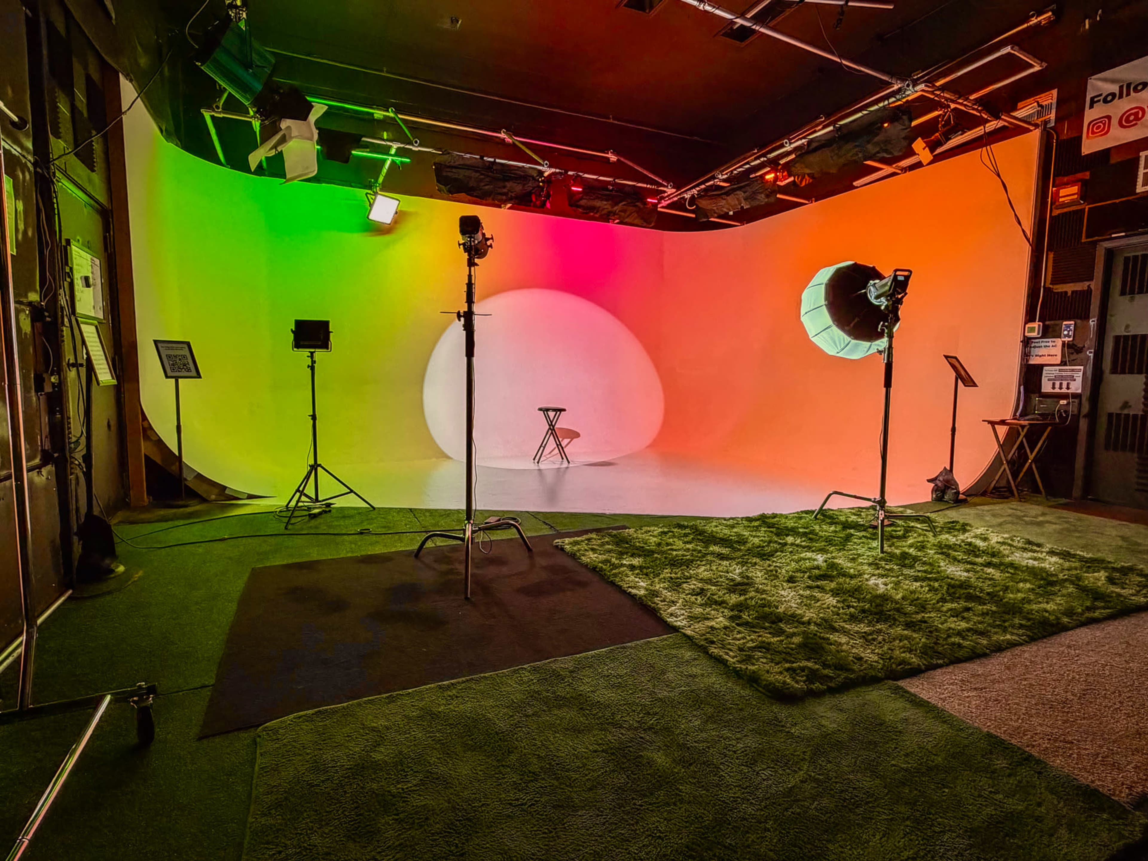 The image shows a photography studio with a curved backdrop featuring colorful lighting, tripods, and a stool positioned in the center on a green carpet.