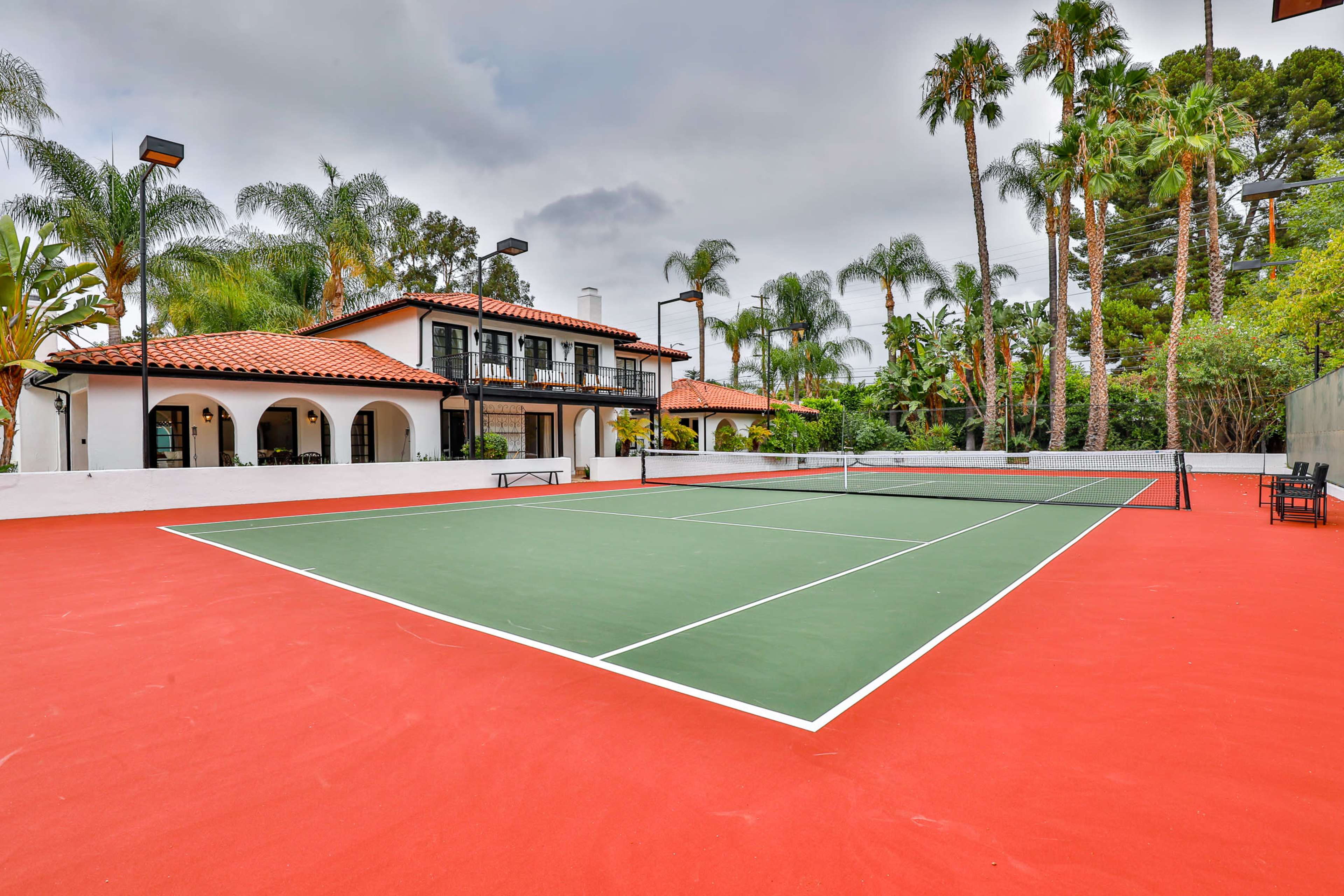 A tennis court with a red surface and green markings is situated in front of a large, two-story house surrounded by palm trees.