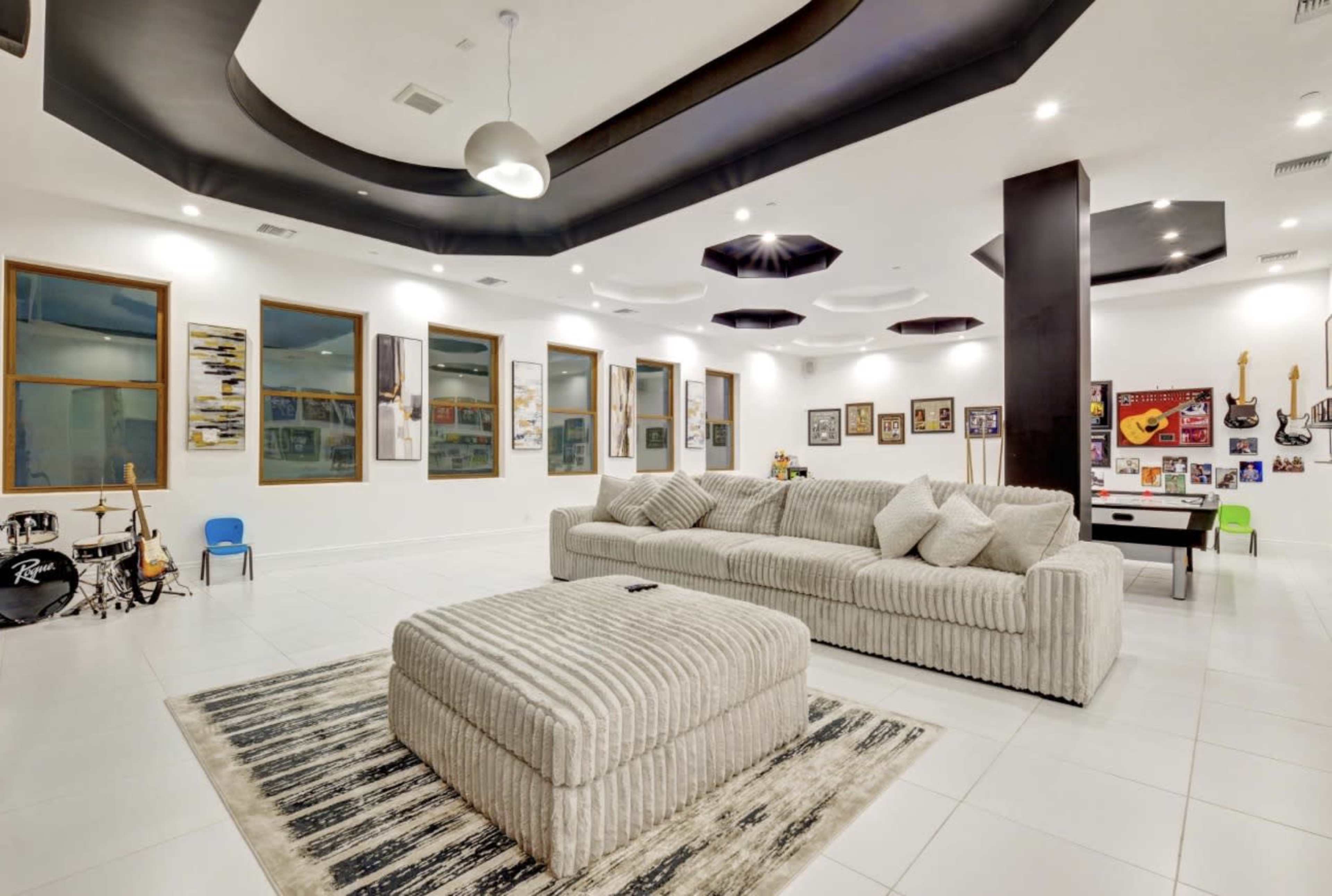 The image shows a modern living room with a striped sectional sofa, a large ottoman, and framed pictures on the walls, complemented by various musical instruments in one corner.