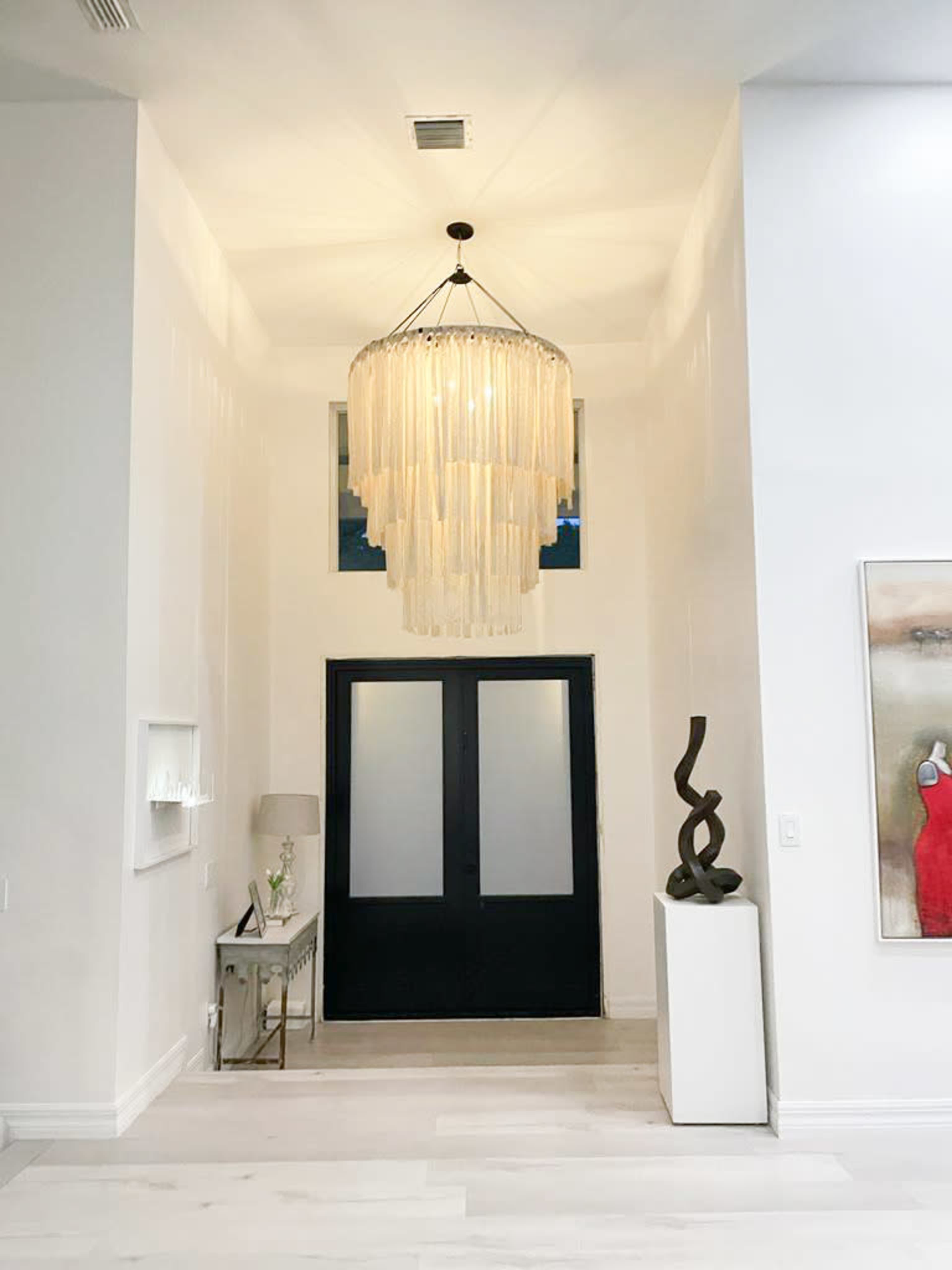A modern entryway featuring a large chandelier, a pair of black double doors, and minimalist decor.