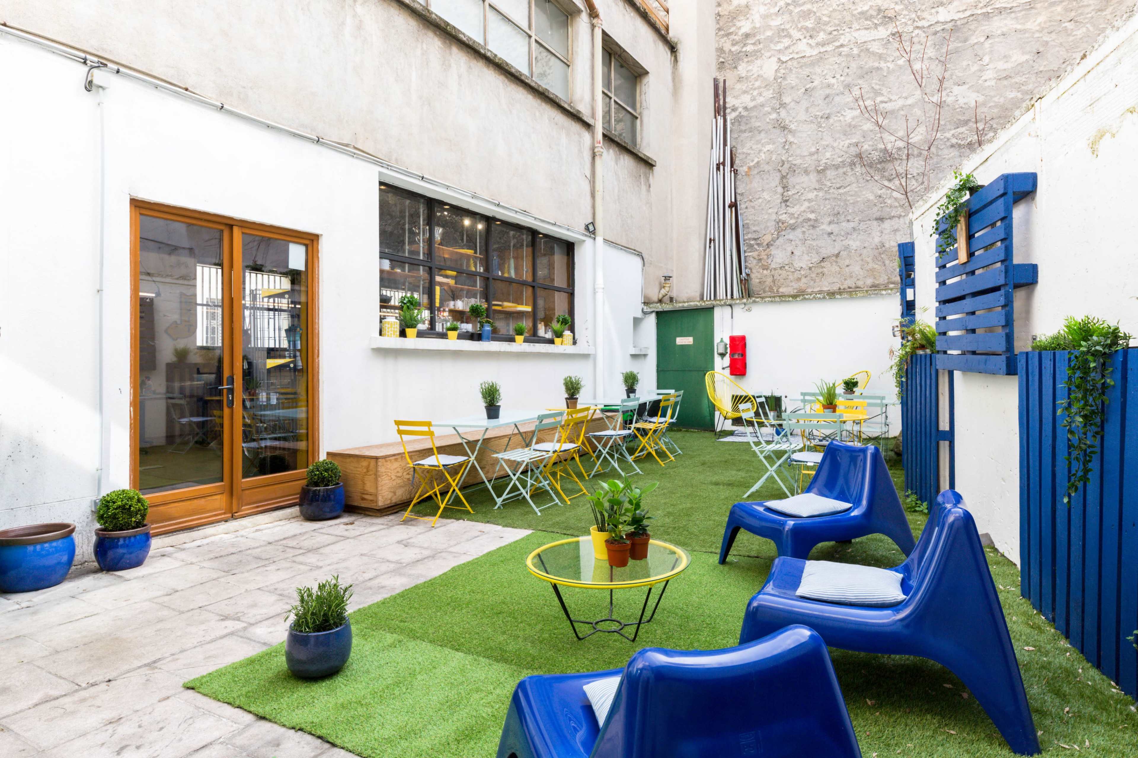 The image shows an indoor-outdoor space with a green lawn, blue chairs, yellow tables, and potted plants, bordered by a beige wall and a wooden door.