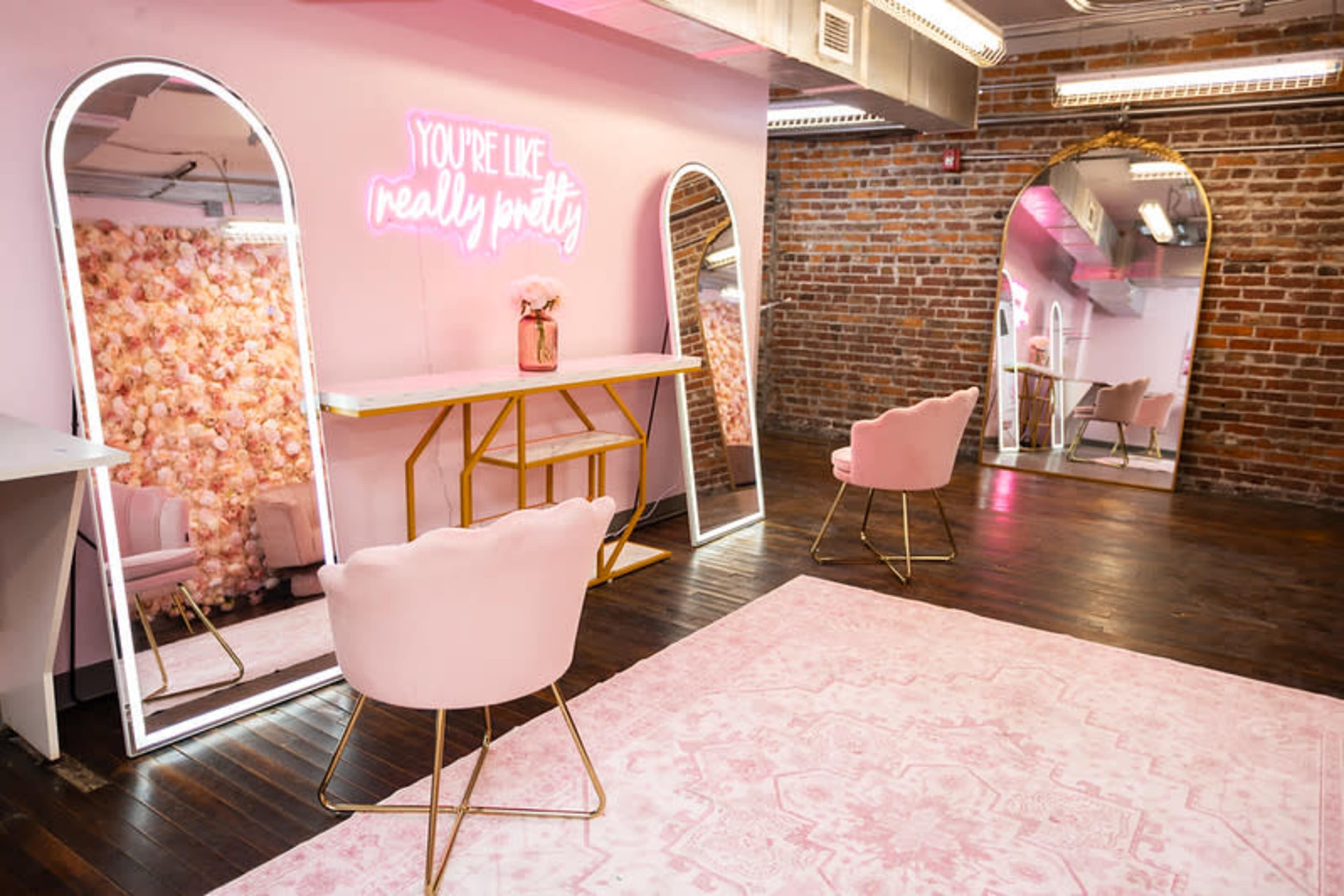 The image shows a bright, stylish room featuring a pink seating area, decorative mirrors, and a neon sign against a brick wall.