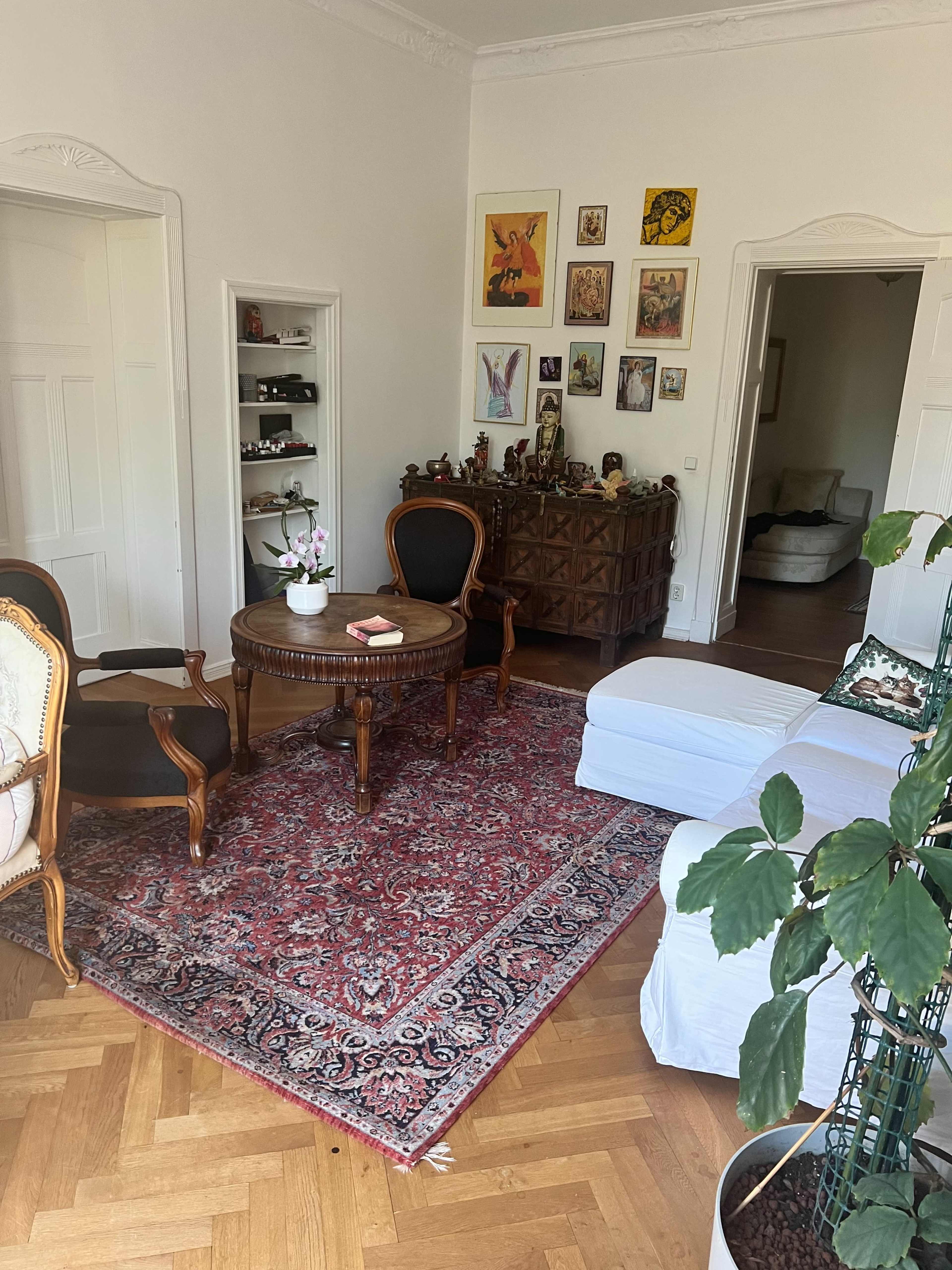 A living room with a round wooden table, two chairs, and a cabinet on one side, featuring a patterned rug and several framed artworks on the walls.